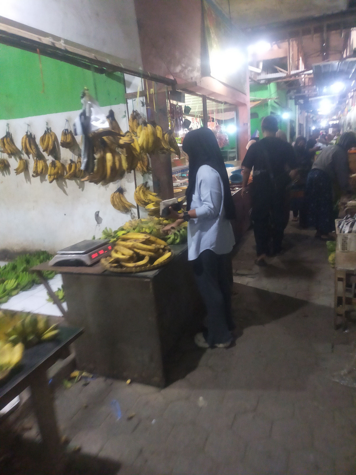 In the city of Trayeman, Indonesia, a market scene unfolds, dominated by the presence of numerous bunches of bananas displayed for sale. The image captures a vibrant, yet somewhat dimly lit, environment. In the foreground, a woman in a light blue top and dark pants, wearing a headscarf, stands near a scale. She appears to be interacting with the bananas or the scale, possibly weighing them for a customer. Behind her, several individuals are visible in the background, suggesting a bustling marketplace.
To the left of the frame, a large quantity of green bananas is laid out on the ground, indicating a fresh supply. Above, bunches of ripe yellow bananas hang from a structure, creating a visually striking display. A digital scale sits on a table in the middle ground, ready for transactions. The background reveals more stalls and structures characteristic of an Indonesian market, with various goods and people present, though many are out of focus. The overall impression is of a busy agricultural market, likely early in the morning or late in the evening, given the lighting conditions. The floor is paved with interlocking bricks, and the walls are painted in muted tones, with a bright green section on the left wall.