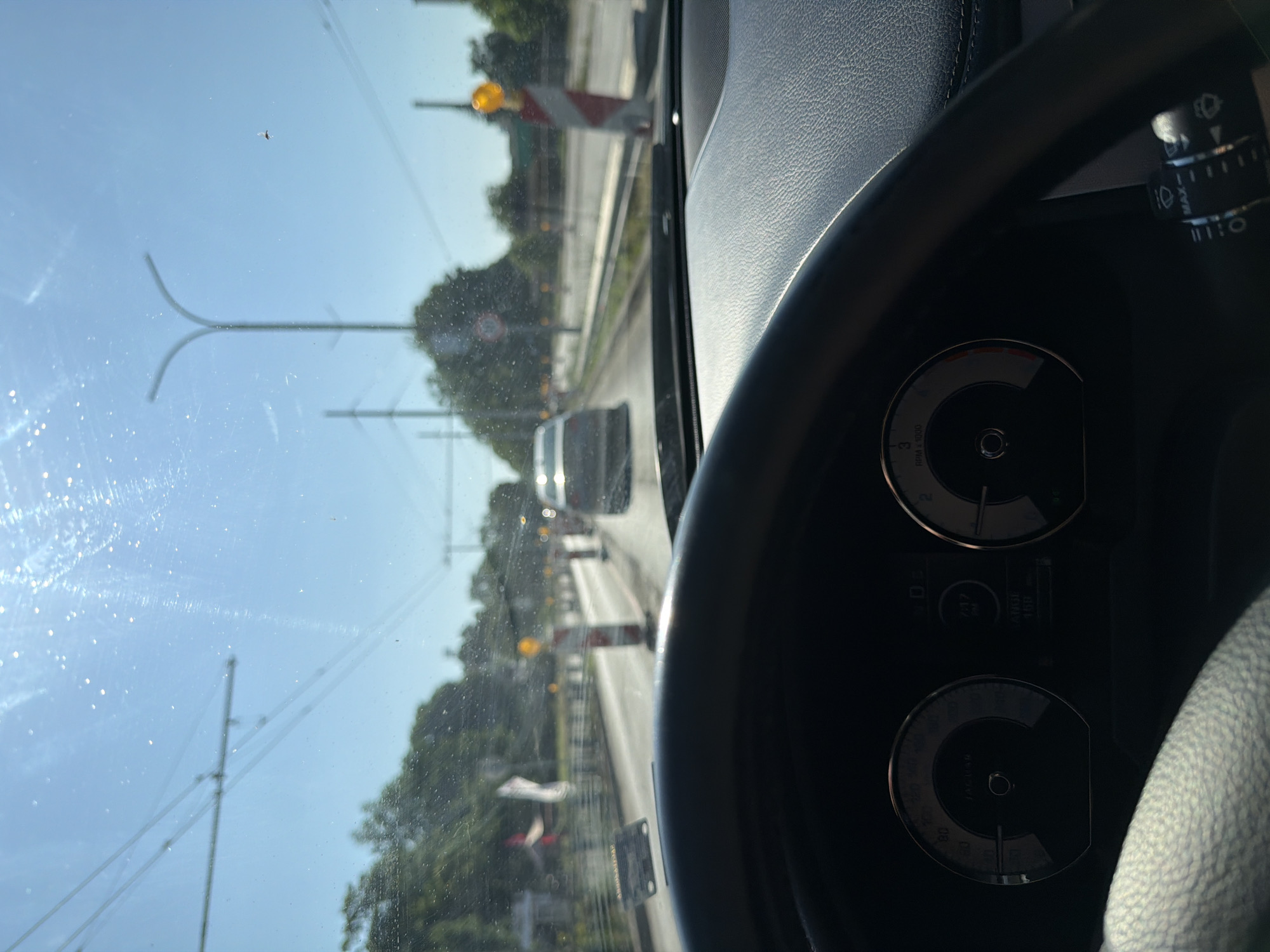 The image is a first-person perspective from inside a car looking forward through the windshield. The view shows a road with some traffic, construction barriers, and trees. The steering wheel and part of the dashboard are visible in the foreground. The sky is bright blue with some streaks of sunlight and what appears to be dirt or dust on the windshield. There are overhead power lines visible. The road appears to be under construction with orange and white striped barriers lining the side. Traffic cones are also present. A car is visible ahead in the right lane, and another car is further ahead in the left lane. The overall scene suggests a sunny day, possibly in a suburban or urban area, with ongoing road work.