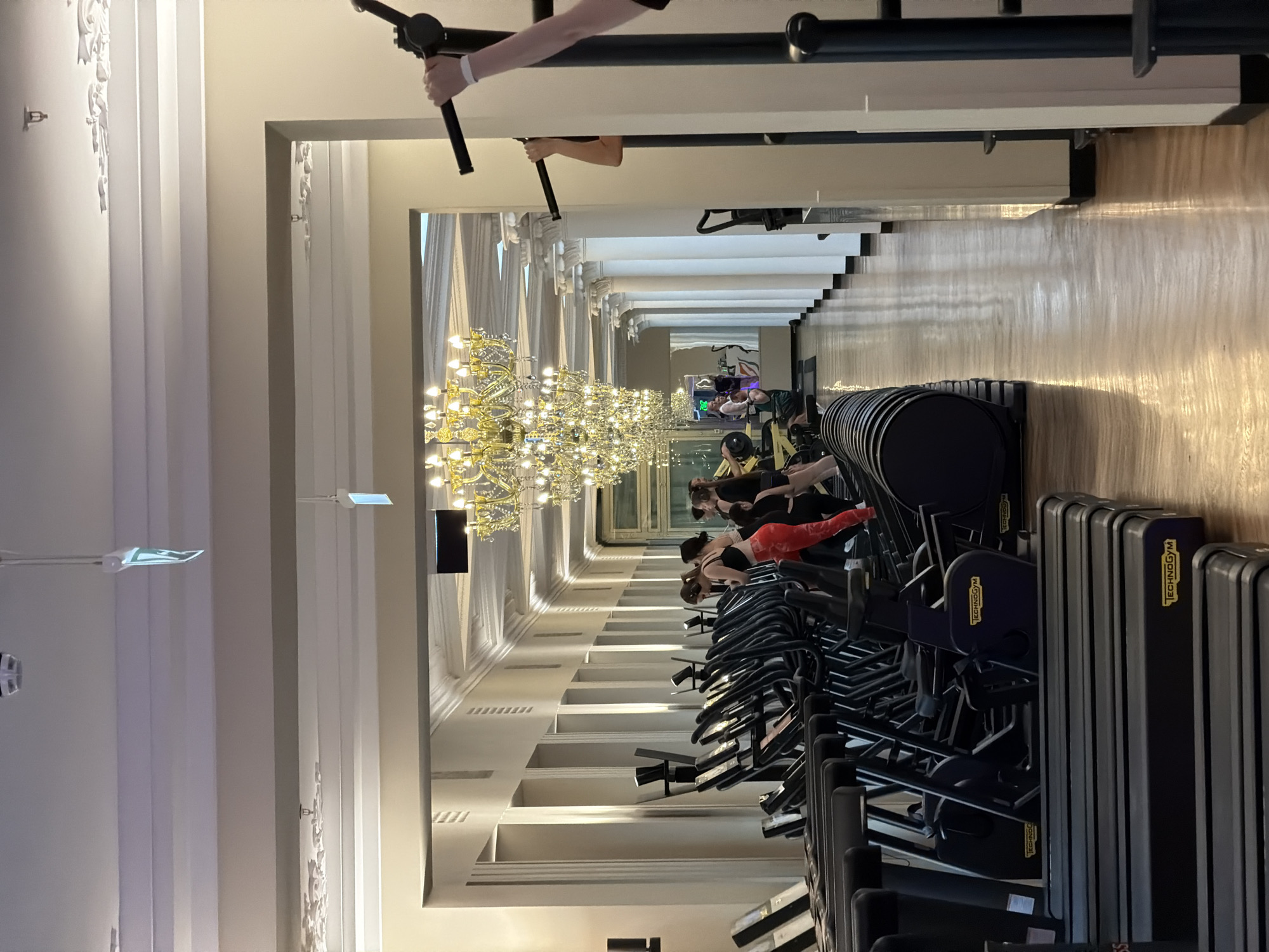 The image depicts a well-lit, opulent gym interior. In the foreground, multiple rows of black treadmills with the "Technogym" logo are visible, suggesting a high-end fitness facility. Further down the corridor, a line of elliptical machines is occupied by several individuals, predominantly women, engaged in their workouts. One woman in the middle is wearing bright red leggings and a black sports bra. The gym features tall, ornate white columns lining the hallway, and a large, glittering crystal chandelier hangs from the ceiling, adding a touch of luxury to the space. The flooring is polished wood, reflecting the ambient light. On the right side of the image, a pull-up bar structure is partially visible, with a person's arm reaching for the bar. The overall impression is of a spacious and elegant gym environment. This gym is located in the historic Nevsky Prospekt area of Saint Petersburg, Russia, within a building that likely dates back to the 19th century, showcasing neoclassical architecture.