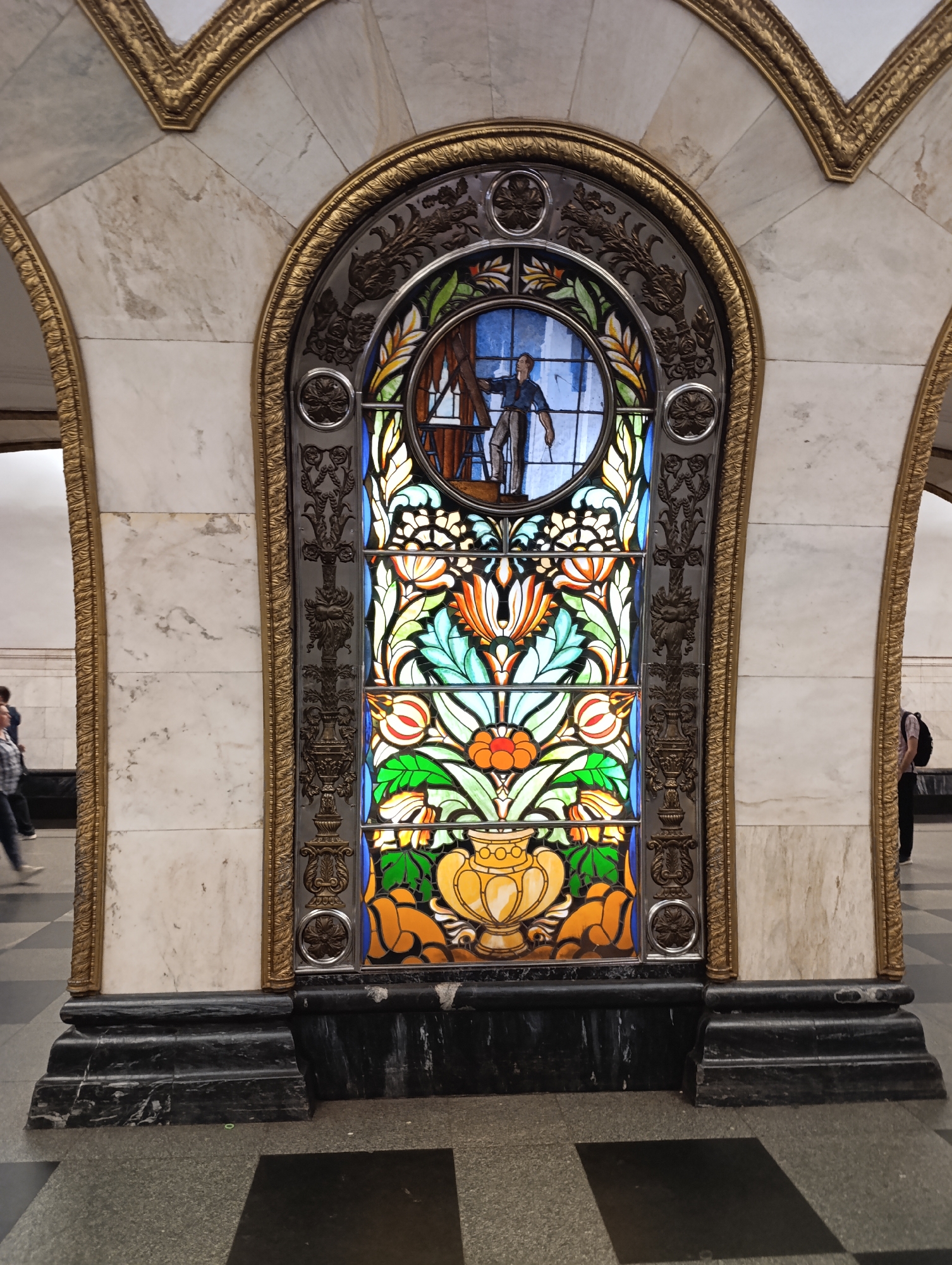 An elaborately decorated arched niche, likely a window or decorative panel, is the central subject of the image. The panel features a vibrant stained glass artwork set within a highly ornate metal frame, which is further enclosed by a golden, intricately carved archway. This archway is integrated into a wall clad with light-colored, veined marble.
The stained glass artwork is vertically oriented. Its upper section is a circular medallion depicting a male figure, possibly a worker or artisan, seen from the waist up, holding what appears to be a tool or perhaps a paint brush in one hand. Below this, the main body of the stained glass is filled with lush, colorful floral and foliage patterns in shades of blue, green, orange, and yellow, culminating in a large, ornate yellow urn or vase at the bottom.
The metal frame surrounding the stained glass is dark-toned and richly detailed with swirling foliate motifs, rosettes, and classical patterns. This frame rests on a multi-tiered, dark stone base that forms the bottom of the niche. The golden trim bordering the marble archway is equally ornate, featuring repetitive, delicate carvings.
In the background, out-of-focus figures of people are visible, some walking, suggesting a bustling public space. More arched structures, similar in style to the main one, extend into the background, hinting at a grand architectural setting. The overall scene conveys a sense of opulent and classical design, characteristic of historical public buildings, such as the Moscow Metro stations known for their lavish interiors. There is no visible text. The lighting suggests an indoor setting, likely artificial.