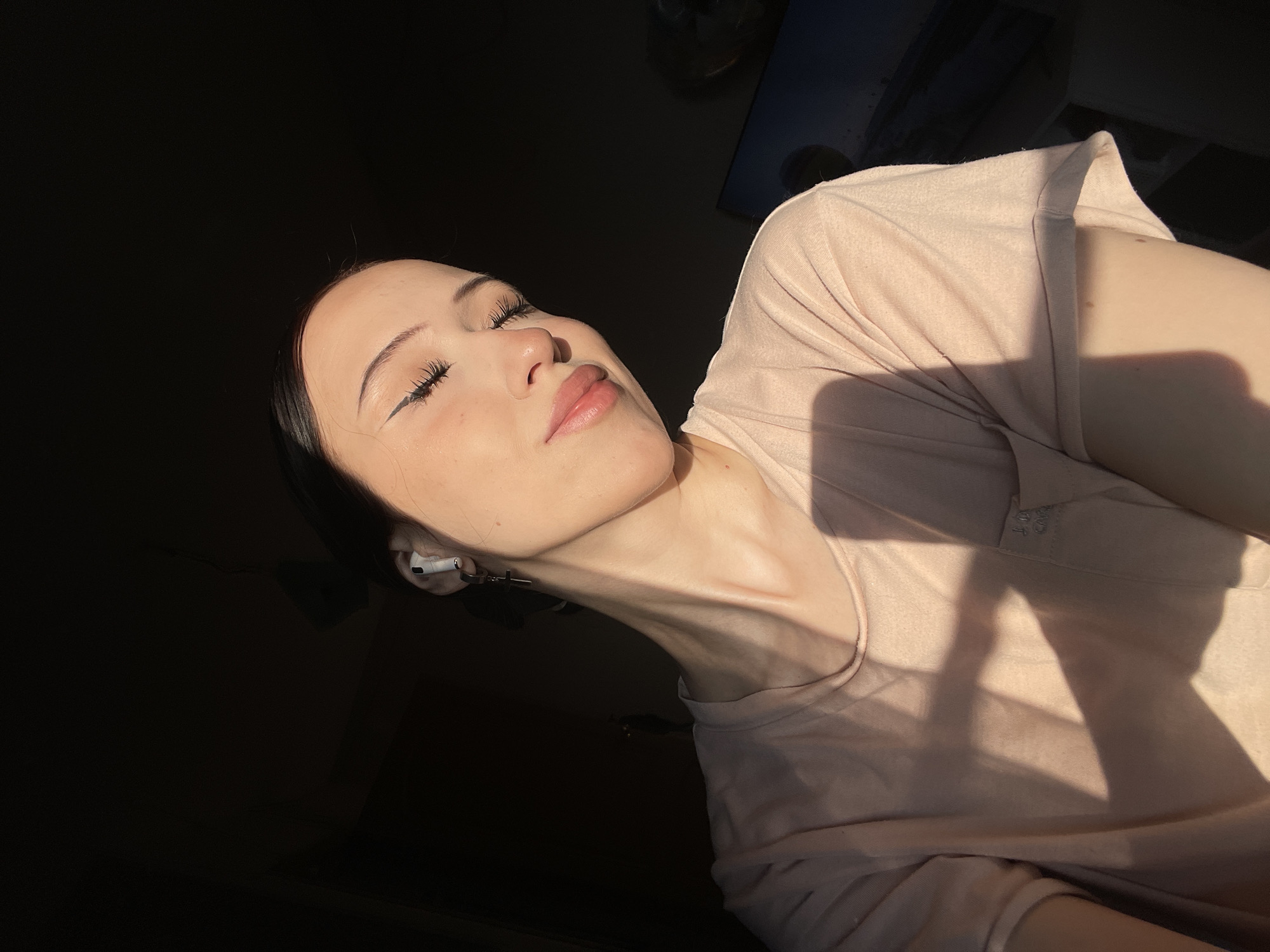 In a dimly lit room, a young woman with her eyes closed and head tilted slightly back, basks in a shaft of warm sunlight. She wears a light pink t-shirt and has a winged eyeliner. An AirPod is visible in her left ear, with a cross-shaped earring attached. Her posture suggests relaxation, perhaps in a moment of quiet contemplation. The sunlight casts a distinct shadow across her t-shirt, indicating the presence of a window or light source. The background is mostly dark, with a faint outline of a monitor or screen visible on the right. The image appears to have been taken indoors, likely in a residential setting. This image was taken in the city of Votkinsk, Russia.