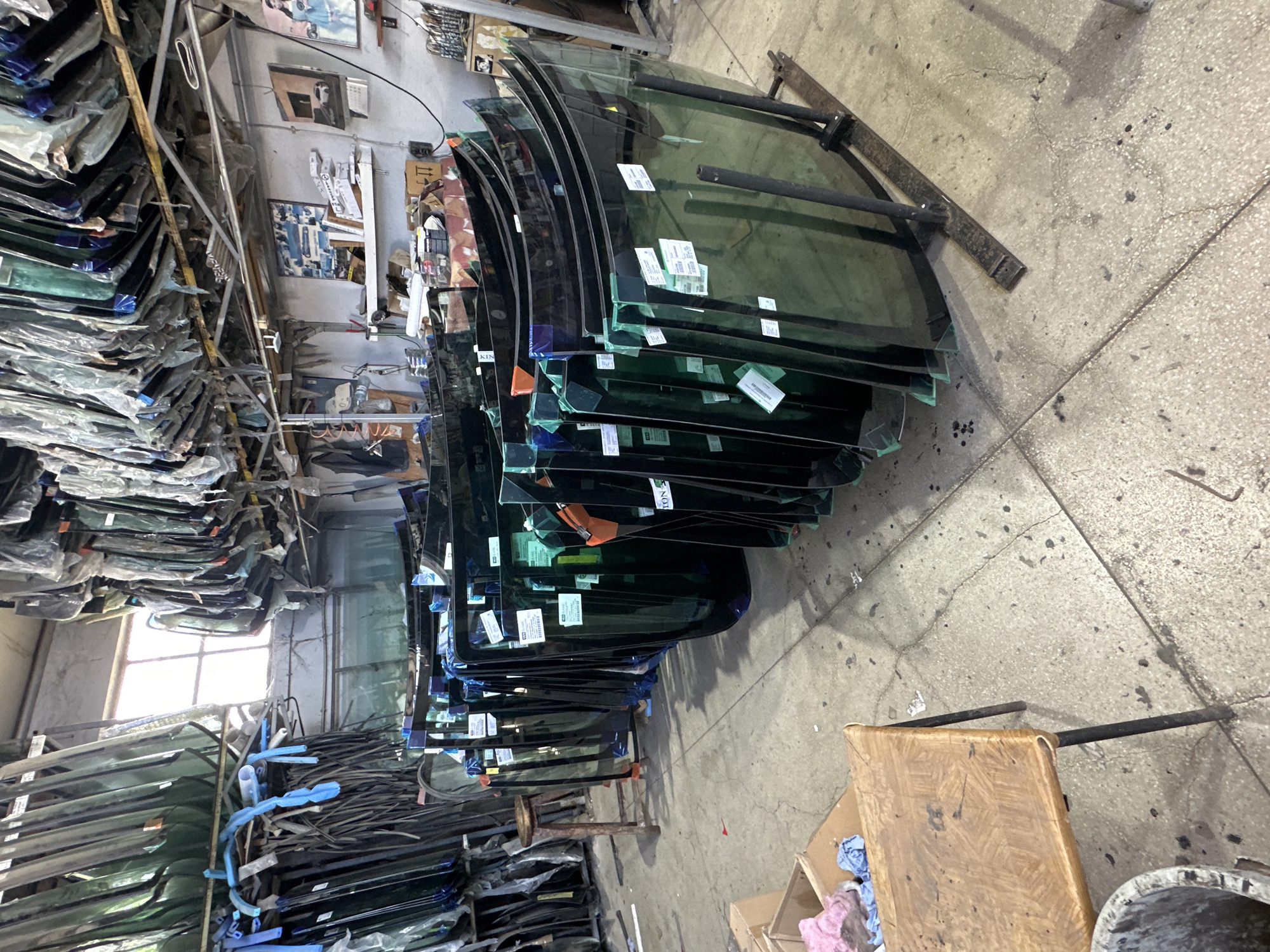 In an indoor automotive repair or storage facility, numerous automotive windshields are displayed, leaning upright on metal racks. The windshields are varied in shape and size, with some appearing to be new and in protective packaging. In the foreground, a low wooden table with metal legs sits on a concrete floor, with a bucket and some scattered rags nearby. In the background, shelves are visible, stocked with various items, and a window allows natural light to enter the space. The overall impression is one of an organized but cluttered workspace dedicated to automotive glass.