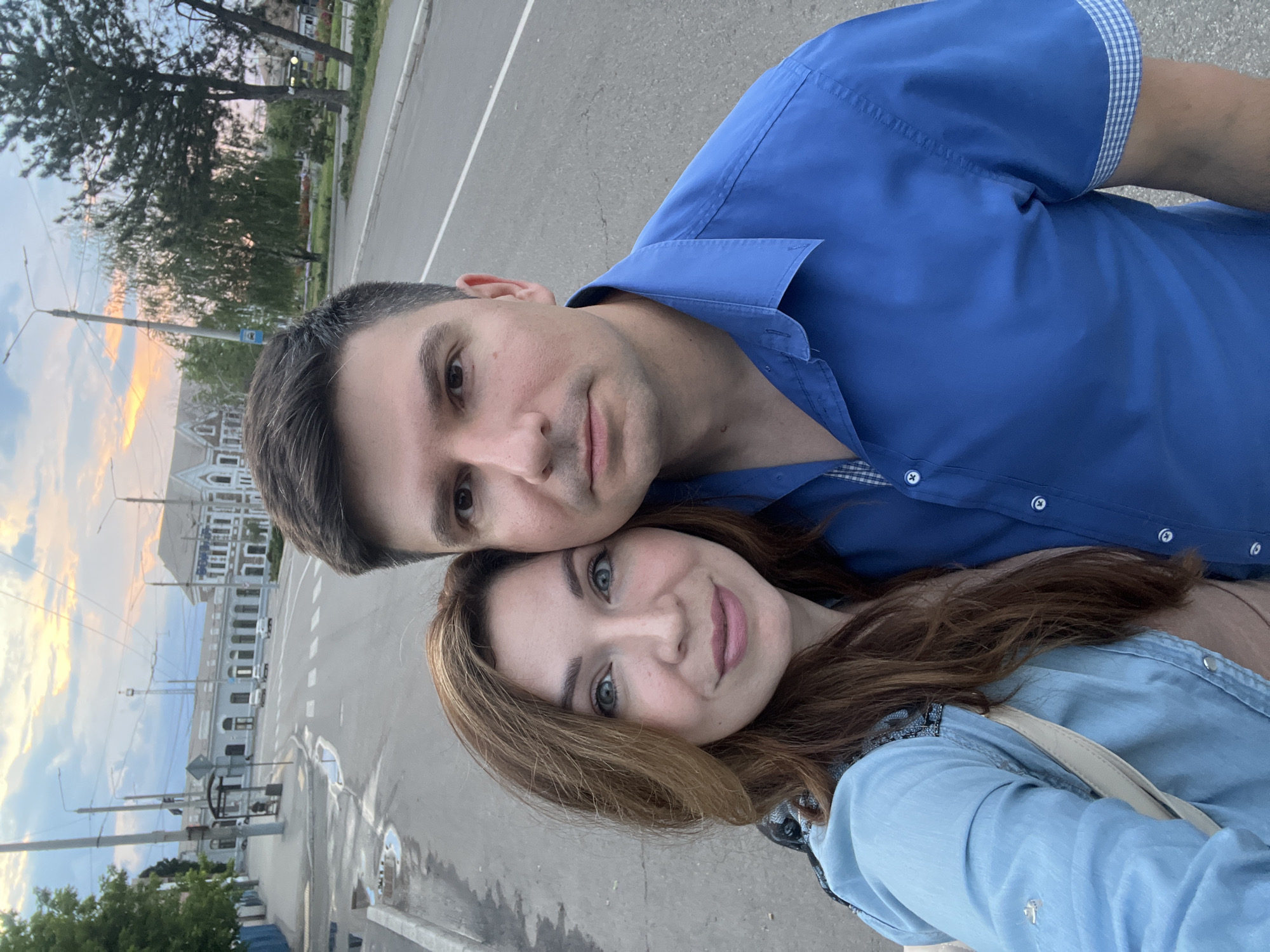 A selfie of a man and a woman standing on a street in Kryvyi Rih, Ukraine, with a train station and trees in the background. The man is on the right, wearing a blue button-up shirt. The woman is on the left, with long brown hair and wearing a light blue shirt. The sky is overcast with hints of sunset colors. The street is paved, and there are buildings and trees lining the sides.