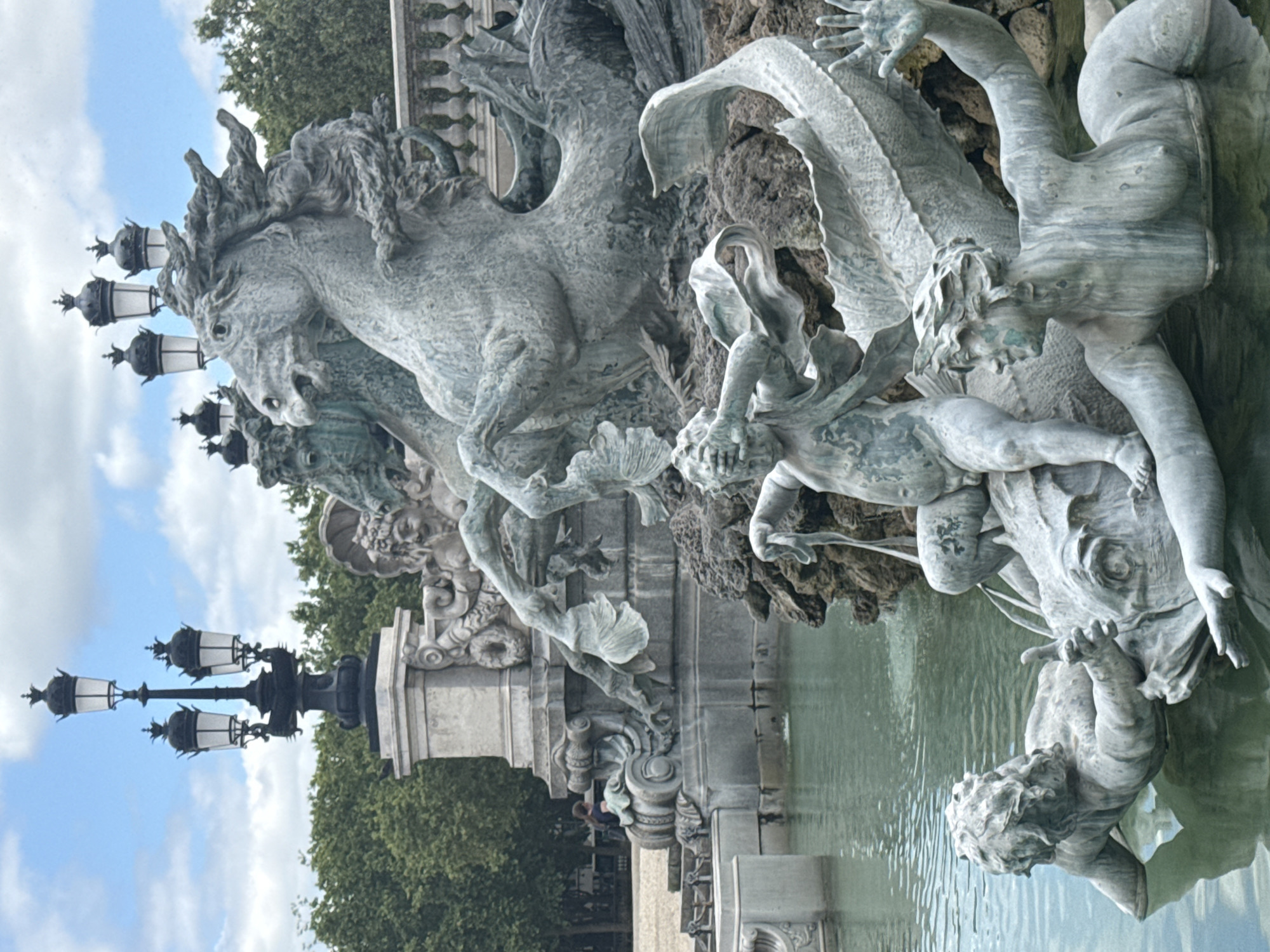 The image shows the monumental fountain of the Place de la Bourse in Bordeaux, France, on a cloudy day. The fountain features several bronze sculptures, including a large sculpture of horses with flowing manes, pulling a chariot, and smaller figures of humans interacting with marine creatures. In the foreground, one human figure is shown partially submerged in water, with a fish-like creature. Another figure, a child-like human, is perched on the fish-like creature, covering its eyes with its hands. A woman's upper body emerges from the water on the right side of the frame, with one arm extended upwards. In the background, ornate lampposts stand against a backdrop of green trees and a blue sky with white clouds. A stone balustrade is visible behind the fountain's sculptures. A person is seated on stone steps on the left side of the image, in front of a large stone monument. The water in the fountain is a greenish hue.