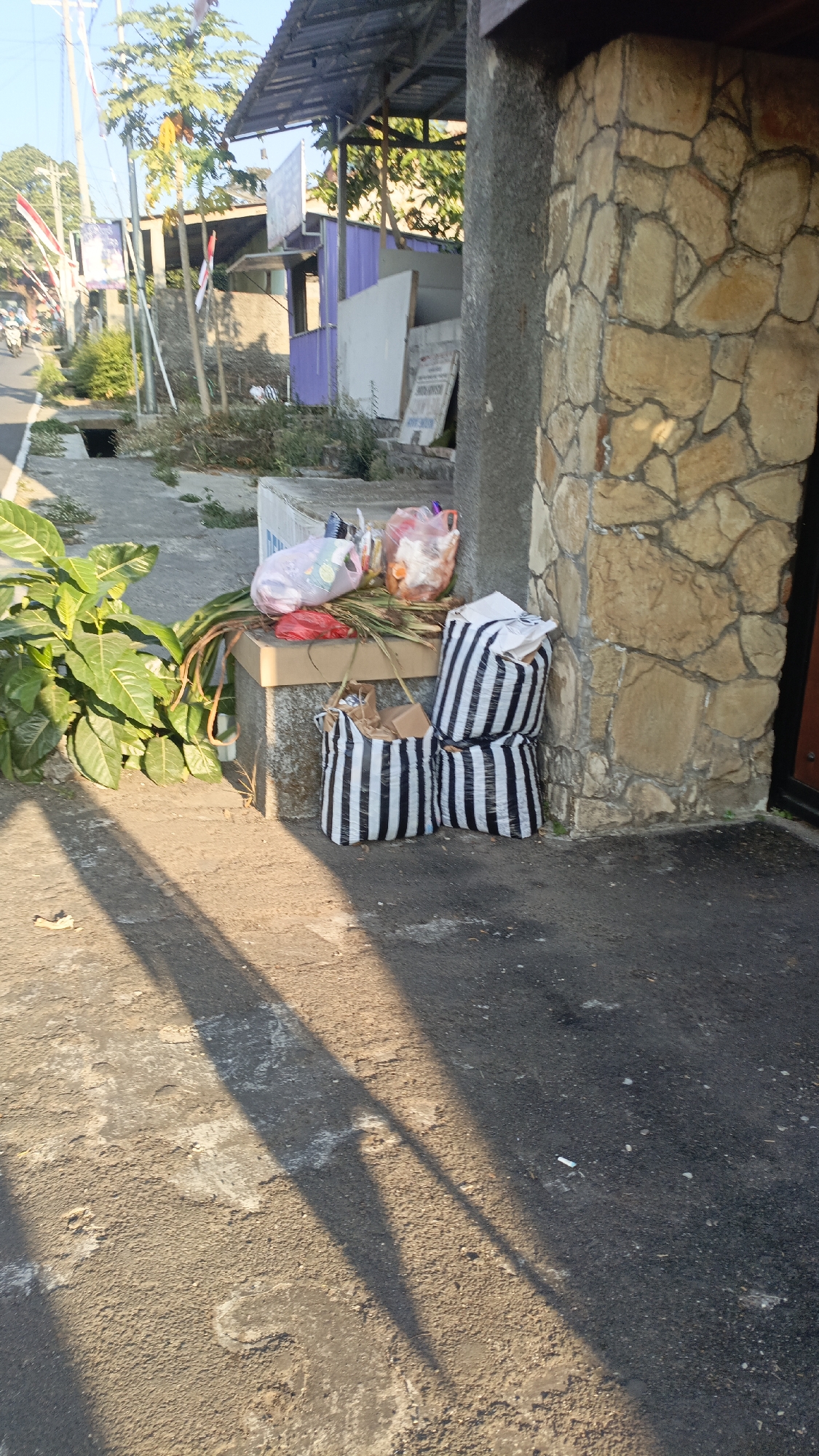 The image depicts a street scene in Salatiga, Indonesia, with a focus on the area next to a building with a stone facade. Several plastic bags, some containing what appears to be groceries or household items, are placed on a raised concrete ledge. Two striped tote bags, one open and containing items, and another closed, are situated on the ground next to the building. A large leafy plant is visible on the left side of the frame. The ground is paved with asphalt, and shadows from overhead structures suggest it is daytime. In the background, there are buildings, trees, and a street with some traffic. The overall scene suggests a residential or commercial area.