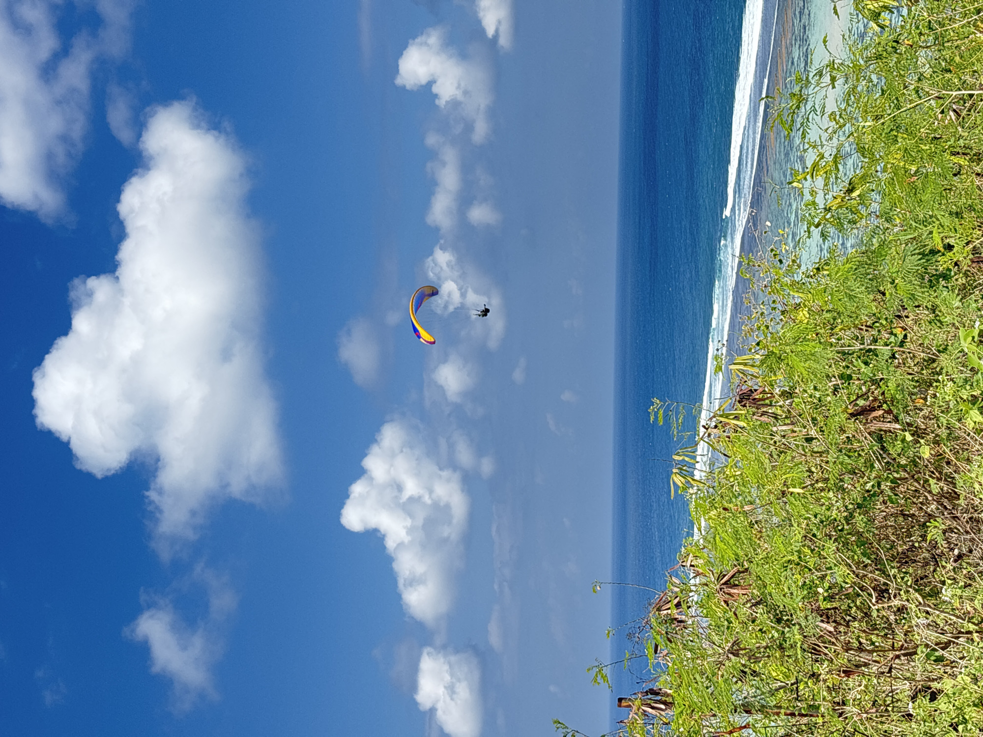 This image depicts a paraglider soaring through a vibrant blue sky dotted with fluffy white clouds, over the ocean near Kuta Selatan, Indonesia. The paraglider, colored in hues of red, yellow, and blue, is suspended in the air. Below, the turquoise waters of the ocean meet a white sand beach, fringed by lush green vegetation in the foreground. The scene captures the essence of adventure and natural beauty, characteristic of the coastal landscapes in Bali.