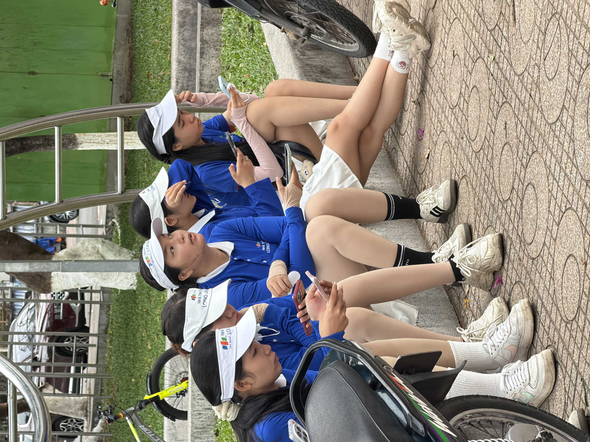 The image depicts a group of young women, likely students or athletes, seated on a concrete ledge outdoors. They are all wearing matching blue long-sleeved shirts with white collars and white visors, suggesting they are part of a team or organization. Several of the women are looking at or holding their mobile phones. One woman on the far left is looking directly at her phone while another next to her is looking away, holding a phone in her hands. The woman in the center is also looking down at her phone, while the woman to her right is holding a phone and looking off to the side. The woman on the far right is looking forward.
The environment appears to be a public space, possibly a park or recreational area, with a paved ground composed of interlocking circular patterns. A portion of a motorcycle is visible on the left side of the frame, and a bicycle with a yellow fork is parked in the background. Beyond the seating area, there is a grassy area with trees and a fence, behind which a dark red car is partially visible. The overall lighting suggests it is daytime. The text on the visors appears to be in Vietnamese, with "NHA THU" and "LONG" clearly legible on some of them. This text, combined with the cultural context of the individuals' attire and the overall setting, suggests the scene could be in Nha Trang, Vietnam, a coastal city known for its beaches and recreational activities.