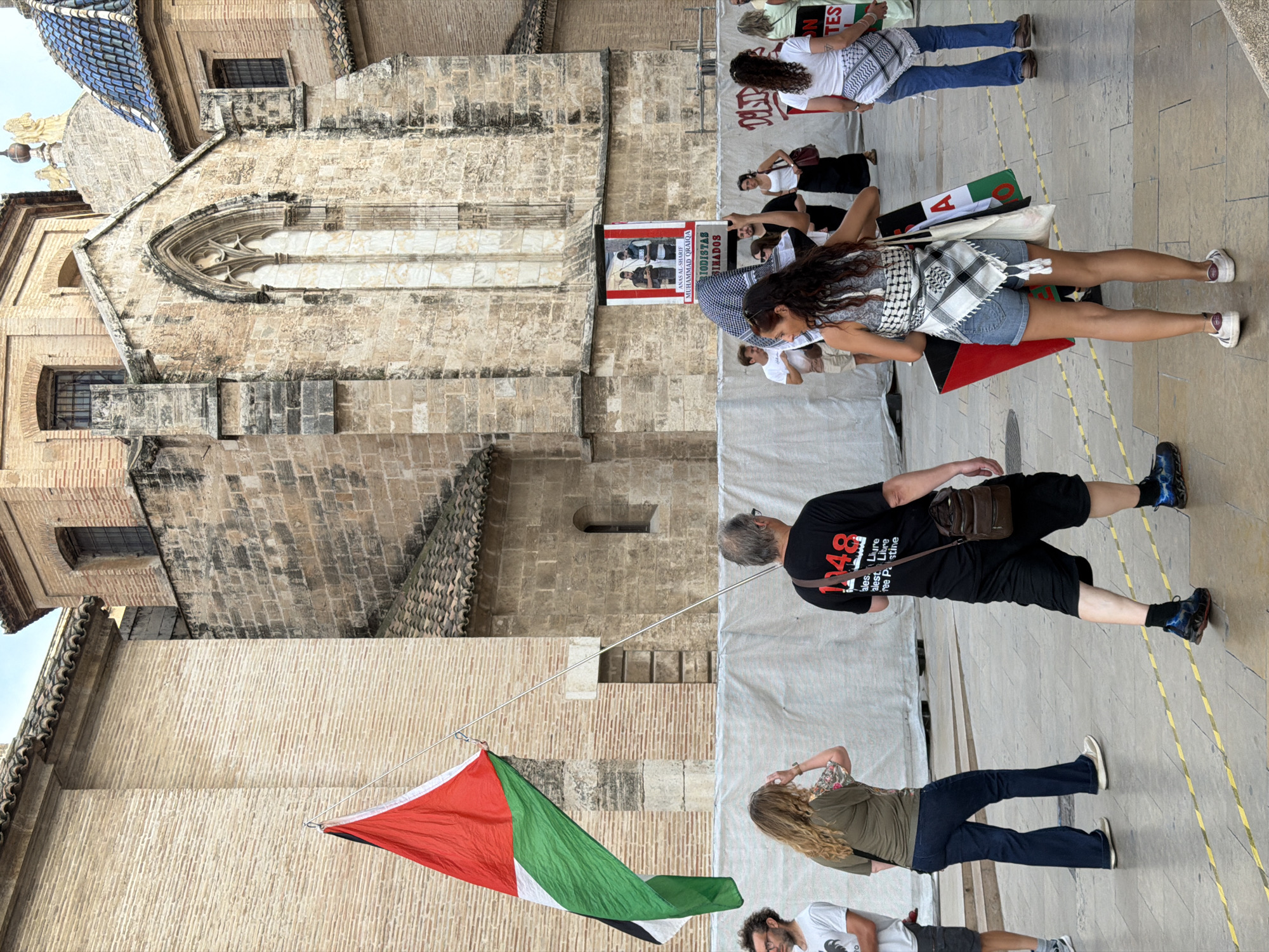 In an outdoor plaza in front of an old stone building, a group of people are gathered. The building features arched windows and a tiled roof. A Palestinian flag, red, white, green, and black, is held aloft by a pole. Several individuals are walking away from the camera, facing towards the building. One man in a black t-shirt with "1948" printed on the back is visible. A woman in denim shorts and a patterned scarf tied around her torso carries a red and black sign. Another woman in a green t-shirt and dark blue jeans walks to the left of the frame. A white banner with text and images is attached to the building in the background. The ground is paved with stone, marked with yellow lines.