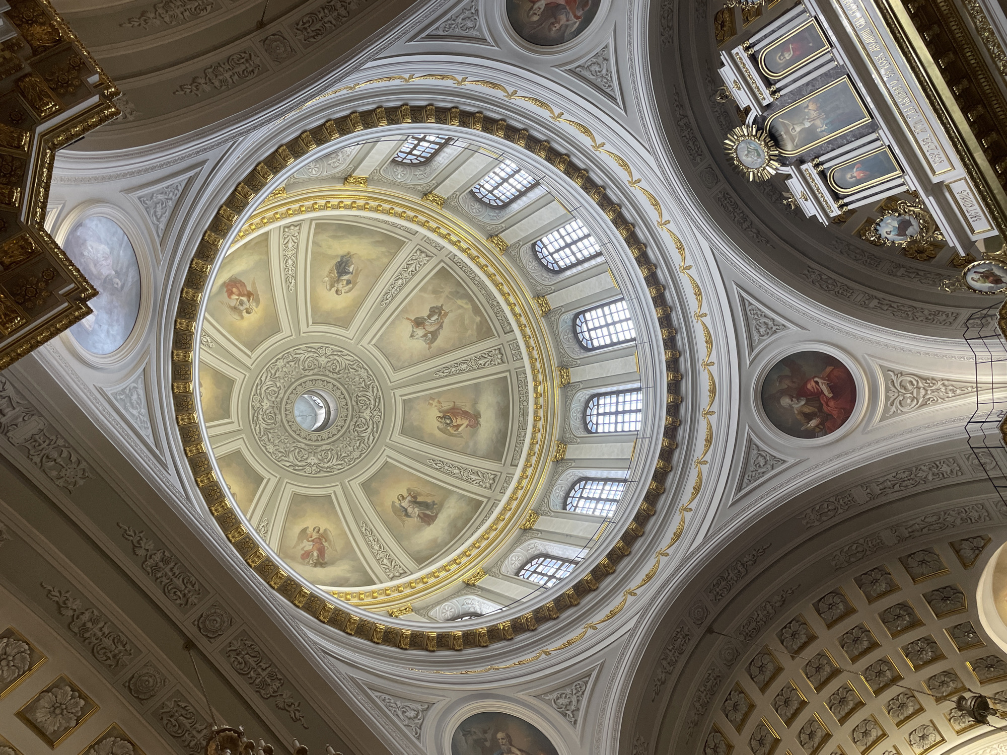The image displays a grand interior ceiling and dome of what appears to be a church or cathedral, viewed from below looking upwards. The central feature is a massive dome, lavishly decorated with frescoes depicting multiple angelic figures—cherubs or angels with wings and flowing drapery—arranged radially around a central oculus that provides natural light. Below this painted section, a series of arched windows, each with multiple panes, forms a ring around the dome, further illuminating the space.
The entire ceiling is adorned with extensive gilding, forming intricate patterns and framing various architectural elements. White or light-colored stucco relief work, featuring detailed floral motifs, scrollwork, and geometric designs, complements the golden accents. Beyond the central dome, smaller circular and oval paintings depicting religious figures or scenes are visible on the surrounding ceiling areas. To the right, a complex, multi-tiered structure, possibly a choir loft or part of the altar, contains rectangular panel paintings, one of which clearly shows a crucifix. A clock face is also visible on this structure.
No specific activity or event is depicted, but the opulent and religious setting suggests a place of worship or a significant historical building, implying a context of religious contemplation or visitation. The scene is brightly lit by natural light entering through the oculus and windows, indicating it is daytime. The grandeur and intricate detailing evoke a sense of awe and reverence.
Visible text in Cyrillic is present on a golden band on the multi-tiered structure to the right. Clearly legible phrases include "ИСУС ХРИСТОС" (Jesus Christ) and "СЪ ВАМИ И ВСЕХЪ" (with you and all).