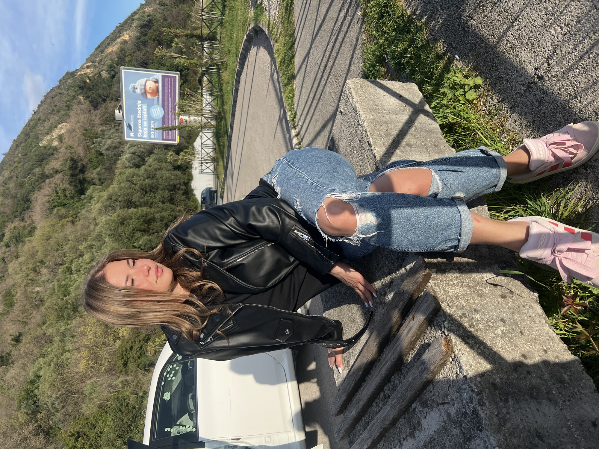 A woman with long brown hair wearing a black leather jacket and ripped blue jeans is lying on a concrete ledge near a white car. She is also wearing pink sneakers. The location appears to be outdoors with a billboard in the background, featuring text written in Serbian. The setting suggests a roadside area with vegetation and a mountain visible in the distance. The photo may be taken in or near Prijevor, Montenegro.