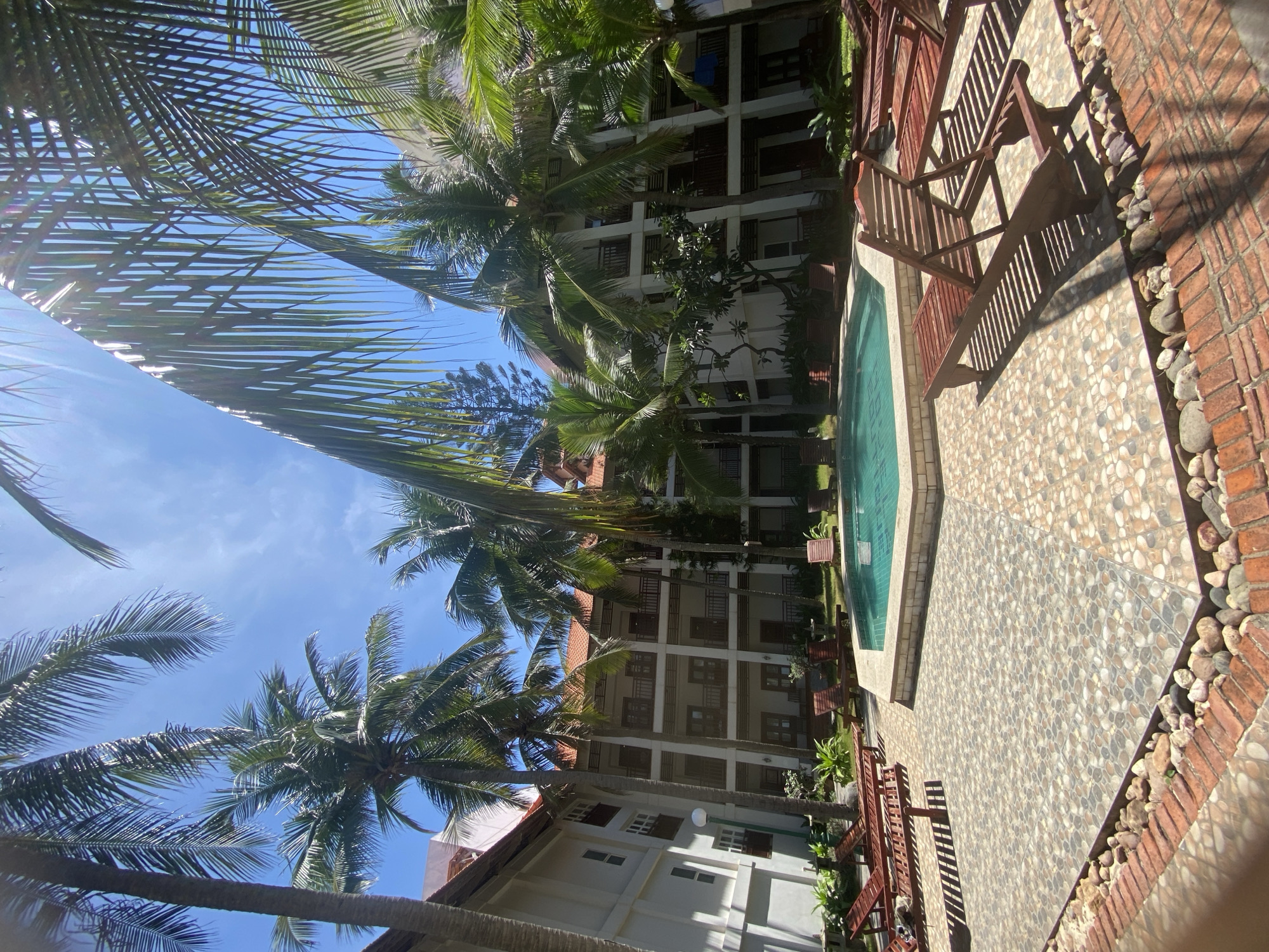 This is a wide-angle, eye-level, outdoor photograph taken in Phu Thuy Ward, Vietnam, showing a resort courtyard with a swimming pool and surrounding buildings. The scene is dominated by lush palm trees casting shadows across the tiled ground and on wooden lounge chairs. In the center of the frame is a hexagonal swimming pool with turquoise water, bordered by a light-colored stone coping. Around the pool, there are several wooden lounge chairs, some unoccupied and some with visible shadows indicating they are empty.
In the background, a multi-story building complex is visible, characterized by white walls, red-tiled roofs, and multiple balconies with dark railings. The architecture suggests a tropical resort or hotel. The sky is a clear, bright blue with some sun glare, indicating it is daytime and likely sunny. The ground is paved with interlocking hexagonal tiles, and along the edge of the paved area, there is a border of red bricks and smooth, rounded river stones. The overall impression is of a peaceful, tropical vacation setting. No people are visible in the image.