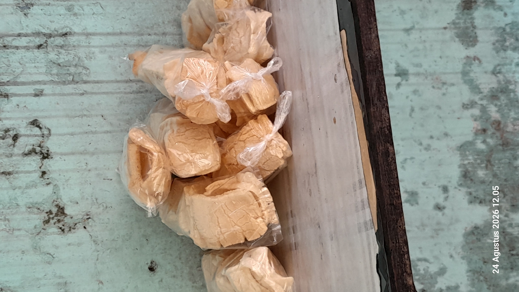 A pile of individually bagged, light brown, porous crackers is stacked on a shelf against a distressed, light blue wall. The crackers appear to be made of a brittle, airy material, with visible cracks and indentations on their surfaces. Each cracker is enclosed in a clear plastic bag, tied shut with a white plastic string. The wall behind the crackers shows signs of wear, with patches of dark, irregular staining and vertical streaks. The shelf itself is a light-colored surface, possibly wood or laminate, with some subtle texture. The bottom portion of the image reveals a dark, textured surface, likely the edge of another shelf or a wooden support structure. In the bottom left corner, a timestamp reads "24 Agustus 2026 12.05". There are no people or animals visible in the scene. The overall impression is that of a storage or display area for a food product in a somewhat aged or rustic environment.