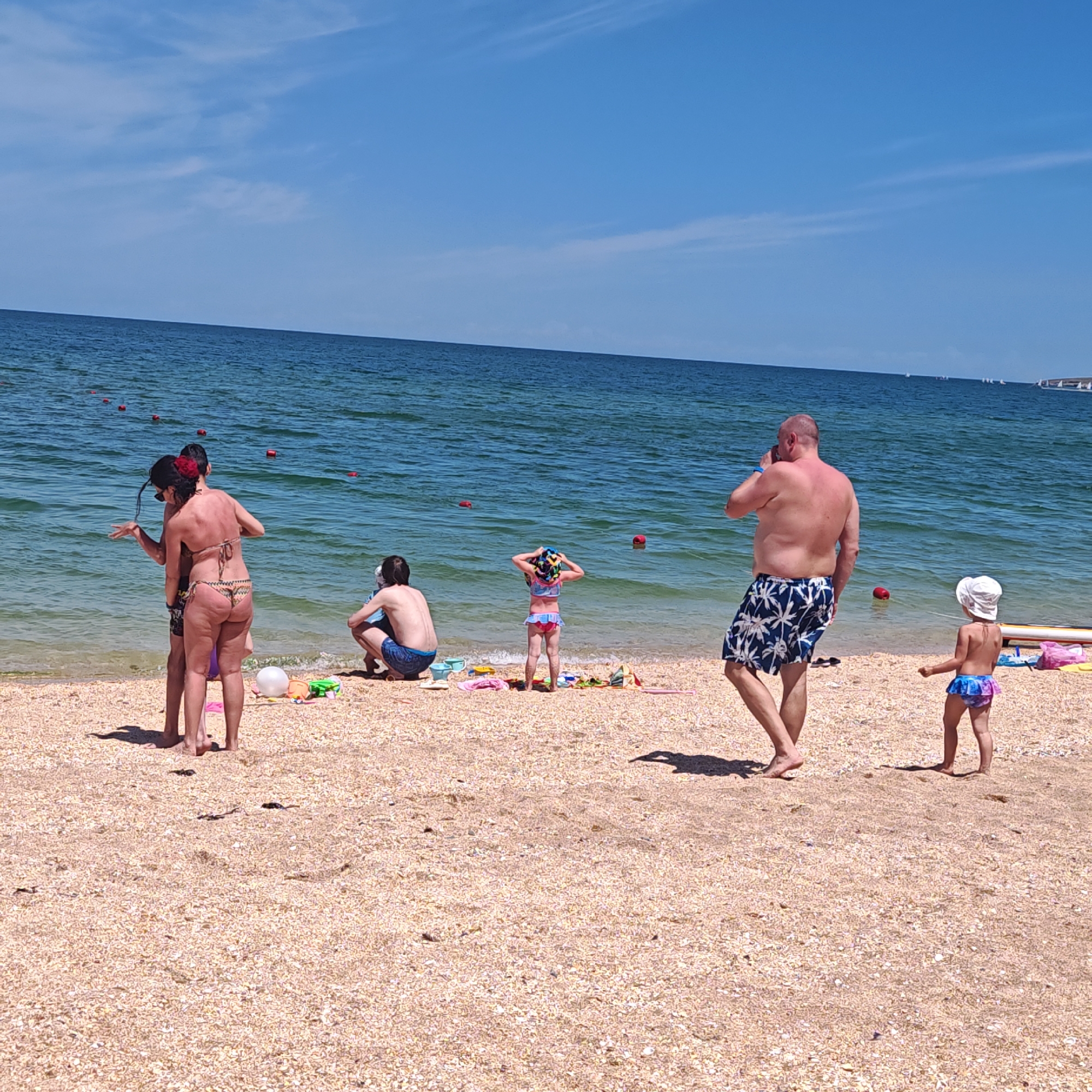 The image depicts a crowded beach scene on a clear, sunny day in Unknown, Unknown. The sandy beach, composed of light-colored sand and numerous small shells, meets calm, blue-green ocean water. The sky is a vibrant blue with scattered wispy white clouds.
In the foreground to mid-ground, several individuals are present. On the left, a woman wearing a patterned bikini with a red flower hair accessory stands facing slightly away from the camera, gesturing towards the water with her left hand, interacting with a partially obscured child or younger person in dark swim shorts. Her back shows evidence of sun exposure. Further into the frame and slightly to the right, a man in blue swim shorts is crouched near the water's edge, facing the sea with his back to the camera. Next to him, a young girl in a colorful two-piece swimsuit and a patterned sun hat stands at the water's edge, also with her back to the camera, looking out at the sea with her hands near her head. Various beach toys and a white inflatable ball are scattered on the sand near these individuals, along with towels or blankets.
On the right side of the frame, a shirtless man with short hair, wearing blue swim shorts with white palm tree patterns, walks away from the water, looking towards his left while holding a phone to his right ear. To his right, a very young child (toddler) in a white sun hat and a colorful two-piece swimsuit stands with her back mostly to the camera, looking down. A light-colored surfboard or paddleboard and a pink bag are visible on the sand behind this child.
A line of red cylindrical buoys extends into the ocean, indicating a marked area. In the far distance, on the horizon, multiple small white sailboats or yachts are visible. The sun casts distinct shadows, suggesting midday conditions.