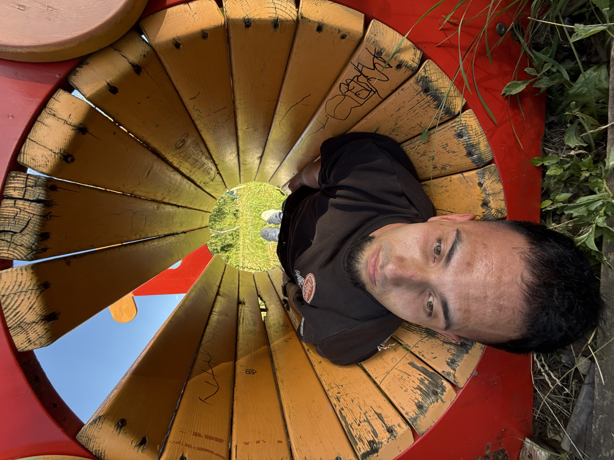 An inverted shot captures a man lying on his back inside a circular wooden structure, possibly part of a playground in Shumen, Bulgaria. The man is wearing a black t-shirt and grey sneakers. The wooden structure is made of curved planks painted yellow and red, with some areas showing signs of wear and graffiti. The background reveals a clear blue sky and green grass, suggesting an outdoor setting.
