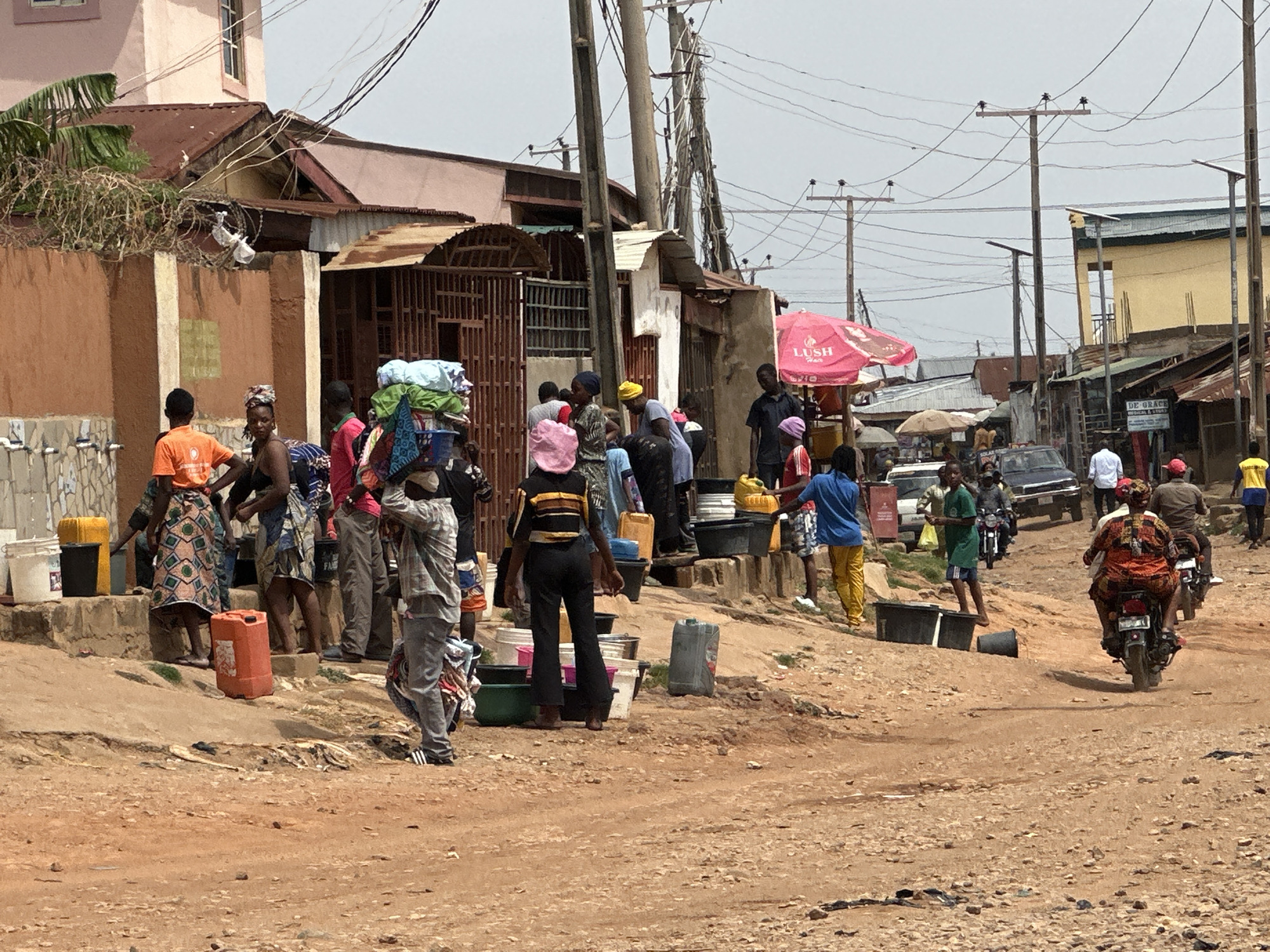 The image depicts a busy street scene in a city, location unknown. A group of people, mostly women, are gathered near a building, many carrying buckets and containers, suggesting a water collection point. Some are carrying items on their heads. The road is unpaved and dusty. Several motorbikes are present, some with passengers, navigating the road. A few cars are also visible in the background. Overhead, numerous power lines crisscross the sky. Buildings in varying states of repair line the street, and a pink umbrella with "LUSH" printed on it is visible.