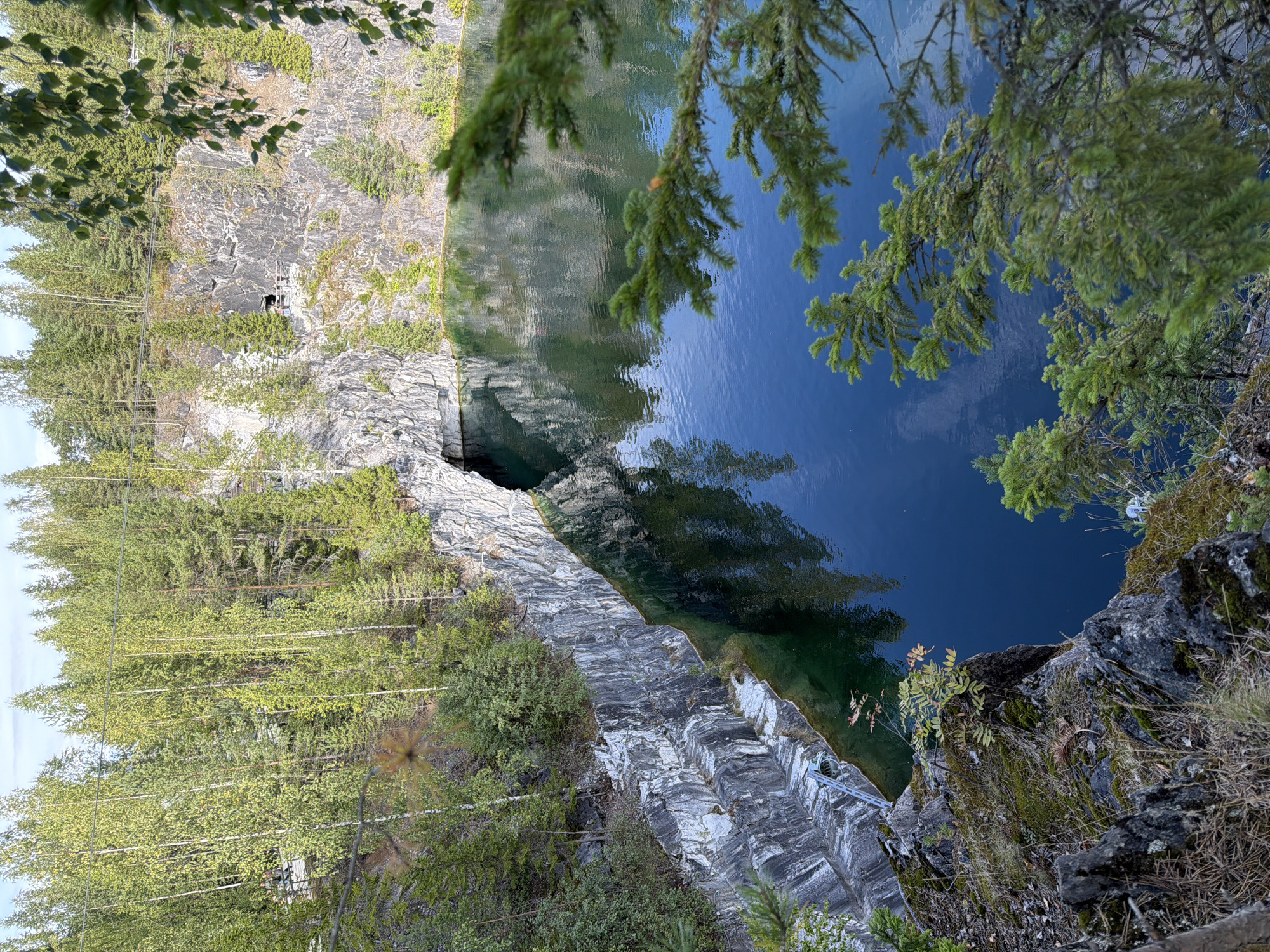 An overhead, outdoor, full shot captures a tranquil, deep blue body of water within a quarry. The water is calm and reflects the surrounding environment, which includes steep, grey, rocky cliffs with visible white striations. Lush green trees and foliage cover the tops of the cliffs and also grow on the rocky outcrops. The scene is bathed in natural daylight, suggesting it is daytime. The water's surface exhibits subtle ripples and its deep blue hue contrasts with the lighter tones of the rock and the vibrant greens of the vegetation. In the distance, on the top of the cliff, a wooden walkway is visible, suggesting a path for visitors.
