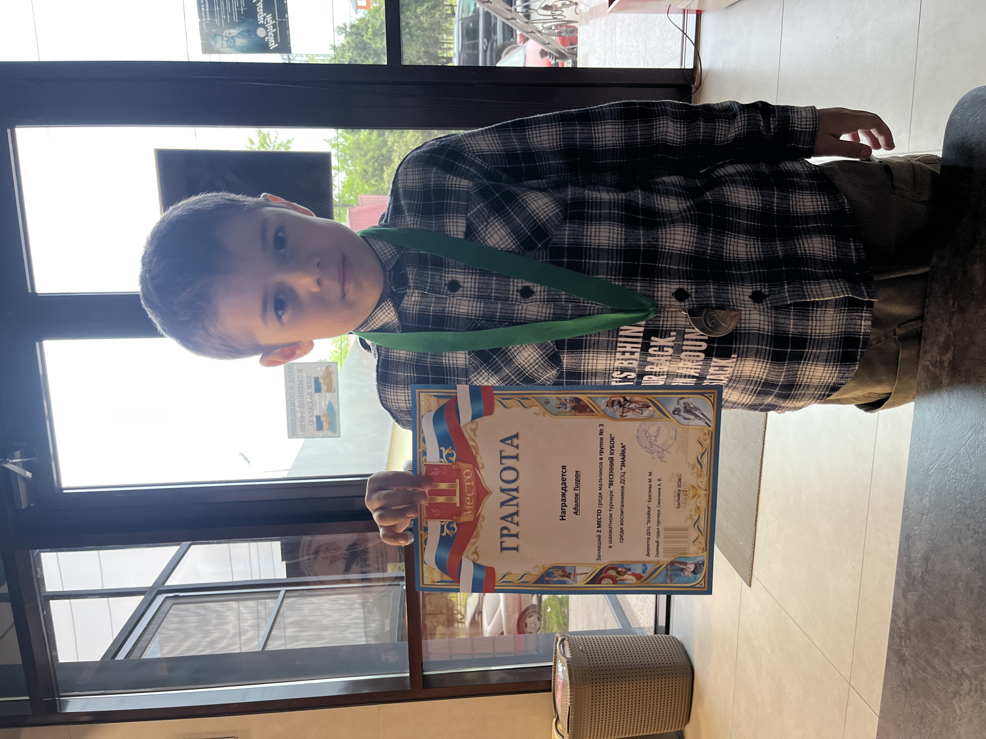 A young boy stands inside a building in Kaspiysk, Russia, holding a certificate ("грамота") that indicates he placed second in the group in the "Spring Cup" chess tournament among students of the "ZNAIKA" center. He wears a green ribbon with a medal around his neck. He is wearing a black and white plaid shirt. The certificate bears the signatures of the Director of the "ZNAIKA" DOC, M.M. Buxaeva, and the Chief Referee of the tournament, A.V. Smagliva. The document states it was issued in Kaspiysk in 2026. The boy is near a window.