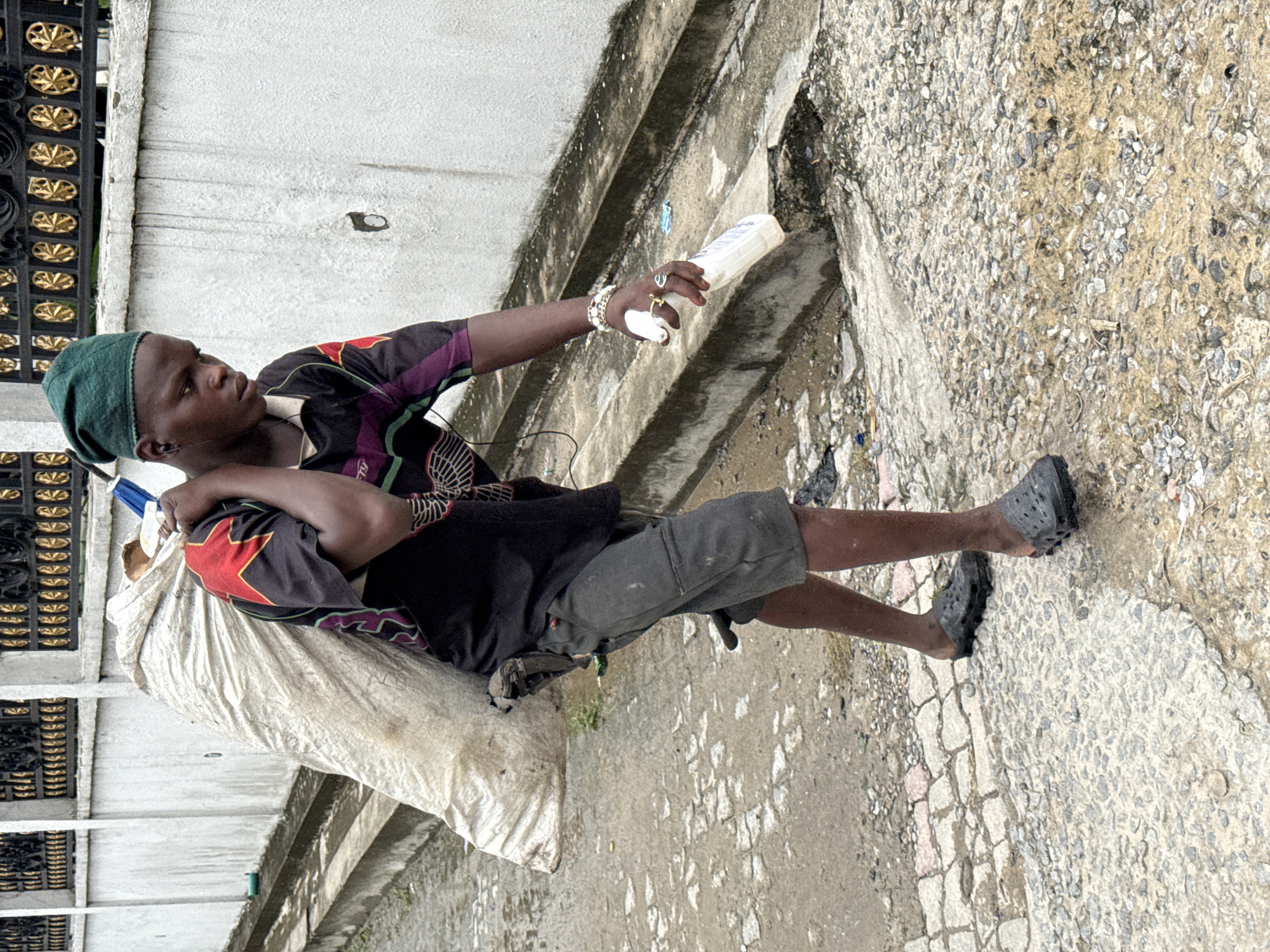 Ado, Nigeria: A young man wearing a green beanie, a black t-shirt with red and purple details, gray shorts, and black clogs carries a large white sack over his shoulder. He extends his right arm, holding a white plastic container. The man wears a bracelet and rings on his right hand. He is walking along a weathered, textured concrete surface adjacent to a wall. Part of a decorative metal fence is visible near the wall.