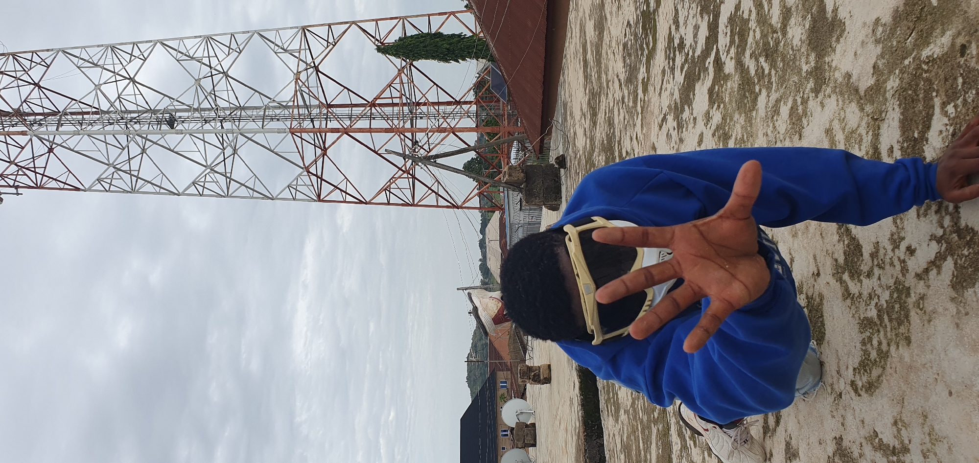 The media file displays a person in the foreground, wearing a blue hoodie and white sneakers. They are positioned on a textured, light-colored surface, possibly a rooftop, with a large, metallic tower structure dominating the background. The tower has a complex lattice design, with sections painted white and reddish-brown. In the distance, several buildings and trees are visible. The sky is overcast with clouds, suggesting a daytime setting with potentially dim lighting. The person is holding their hand out towards the camera, palm facing forward, in a gesture that could be interpreted as a greeting or a request to stop. Their face is partially obscured by large, white-framed glasses. The overall scene appears to be outdoors, in a somewhat industrial or urban environment, given the presence of the tower and buildings. The location is identified as Anguldi, Nigeria. There is no visible text in the image.
