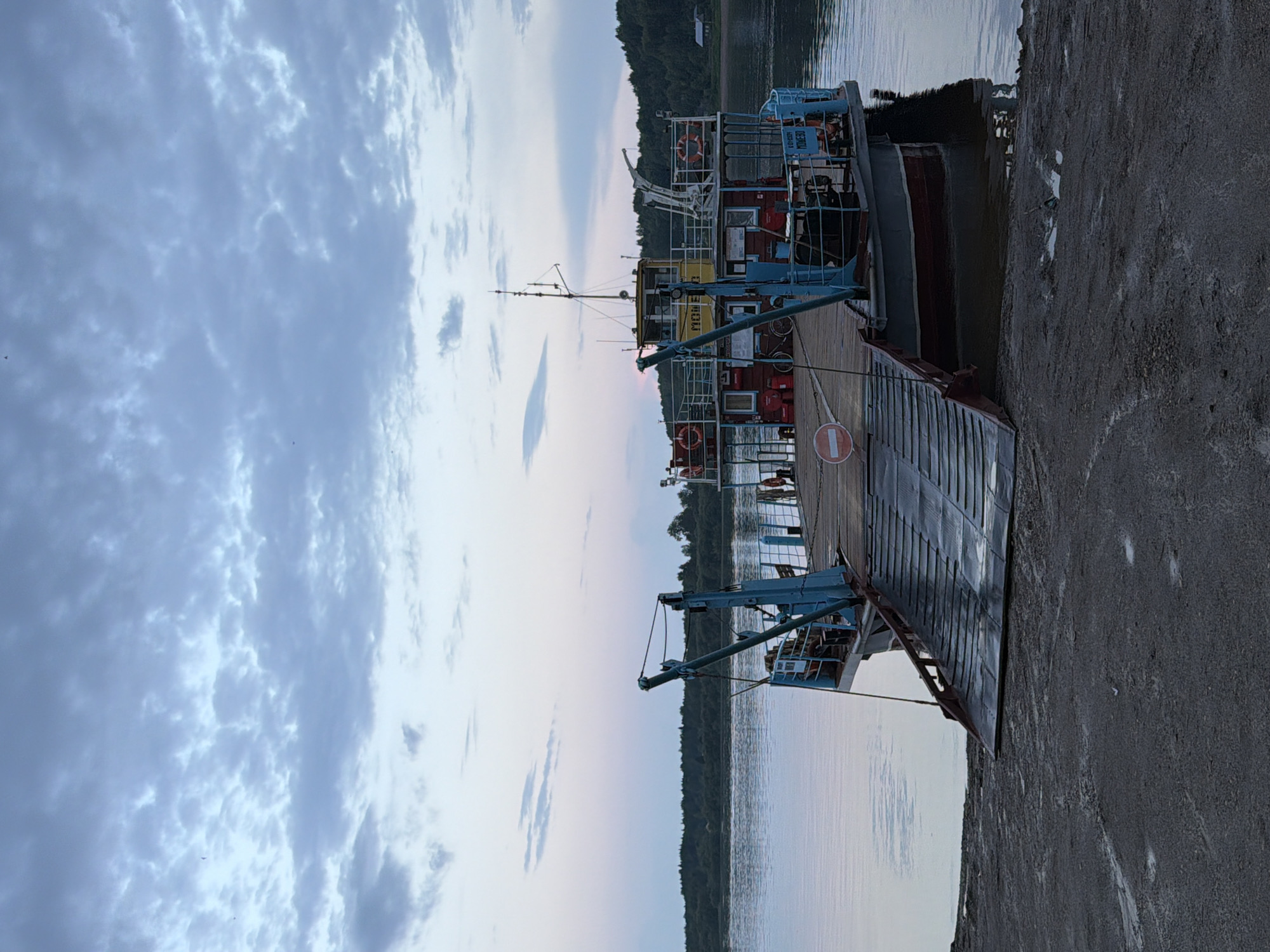 A ferryboat is docked at the shore of a river in Tyulkino, Russia. The ferryboat has a blue ramp that extends from its deck to the shore. The ferryboat has a red hull with a blue superstructure. The superstructure is yellow and has windows. The ferryboat has a mast with antennas. There are life preservers on the sides of the ferryboat. A bicycle is visible on the deck of the ferryboat. The sky is cloudy and the water is calm. Trees line the opposite bank of the river. The ground in the foreground is dark and wet.