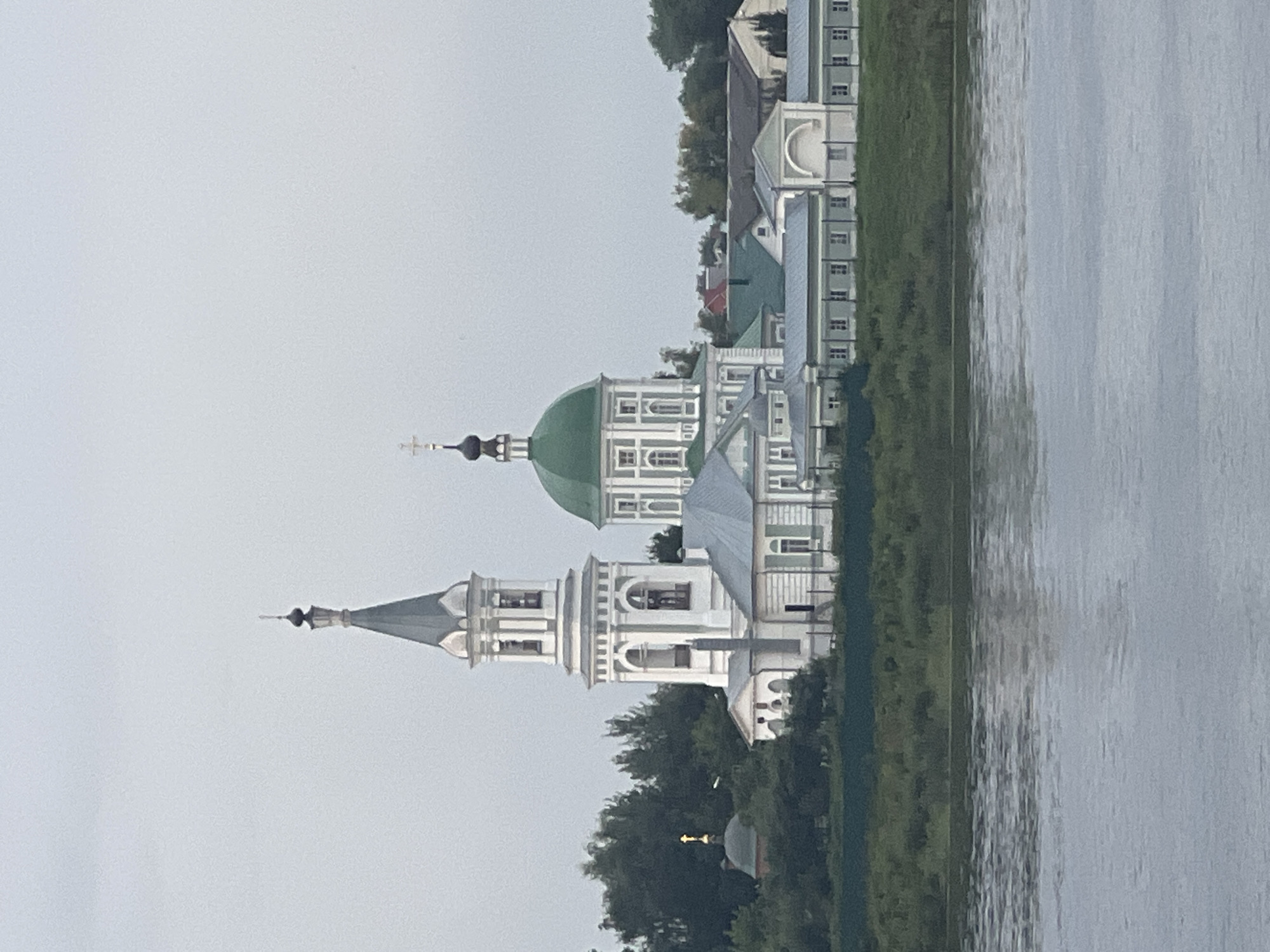 The image shows a panoramic view of a monastic complex on the bank of the Volga River, likely in Tver, Russia. The complex features a prominent white bell tower with a grey spire, topped with a dark onion dome and a cross. Adjacent to the bell tower is a larger building with a striking green onion dome, also crowned with a cross. The buildings are white with grey roofs and green accents, characteristic of Russian Orthodox architecture. Surrounding the complex is lush green foliage, and the foreground is dominated by the grey, rippling water of the Volga River. The sky is overcast and a pale grey, suggesting a cloudy or early morning/late evening setting. There are no people visible in the image. The overall impression is one of serene, historical architecture set against a natural landscape.