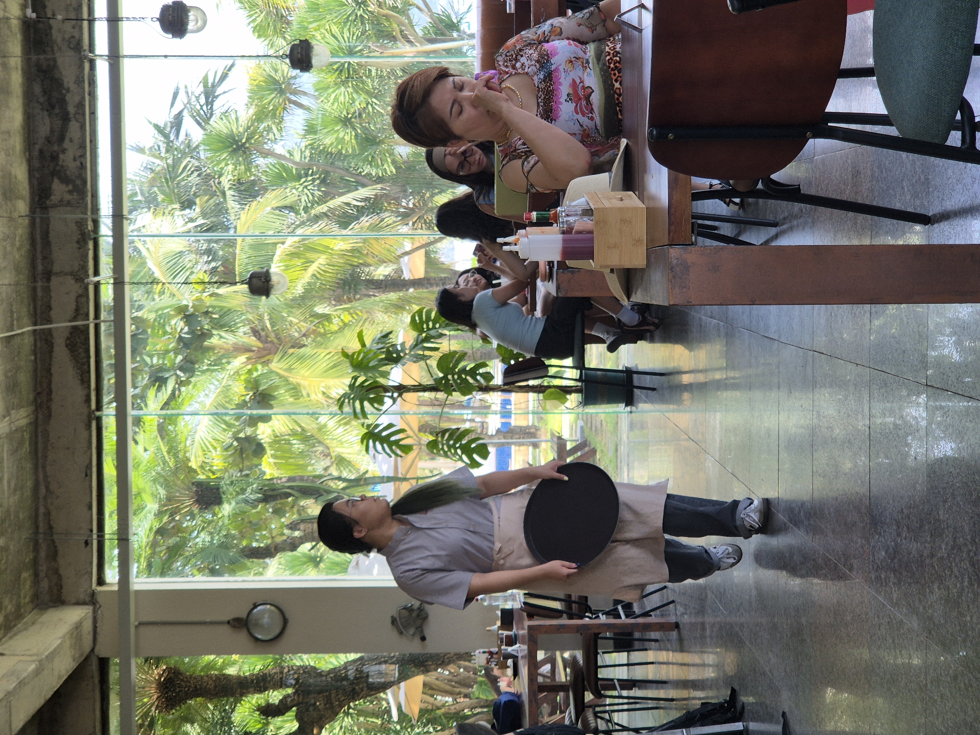 The image captures a moment inside a restaurant in Đà Nẵng, Vietnam, with a large window offering a view of lush tropical foliage. A female server, wearing a grey shirt and a light-colored apron, walks across the polished floor, carrying a black tray. She has dark hair tied in a ponytail and is looking to her right. In the foreground, a woman with short brown hair is seated at a table, her eyes closed as she touches her face. She is wearing a colorful floral and leopard print top. Several other individuals are visible at tables in the background, appearing engaged in conversation or dining. The restaurant interior features wooden tables and chairs, and the overall atmosphere suggests a casual dining setting bathed in natural light.