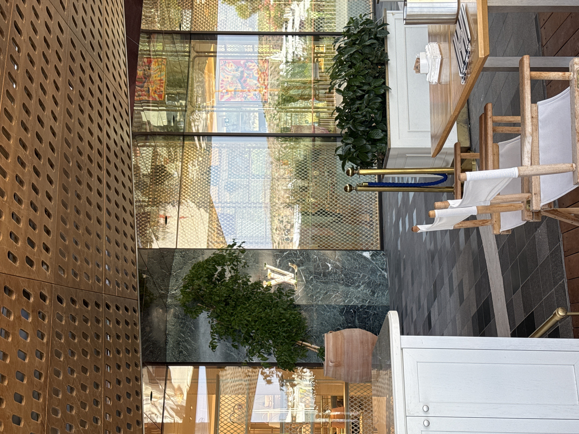 The image depicts an outdoor seating area in front of a Louis Vuitton store. The store features a facade of green marble with a prominent gold Louis Vuitton logo. A large potted ficus tree stands to the left of the entrance. The outdoor area has a wooden deck with a white cabinet on the left and a wooden table with white canvas chairs on the right. The table is set with a menu and what appears to be sugar packets. In the background, a reflective glass building with intricate metalwork is visible, reflecting the surrounding landscape and artwork. The scene is set during the daytime, with clear sunlight casting shadows. The location is identified as Vouliagmeni Municipal Unit, Greece.