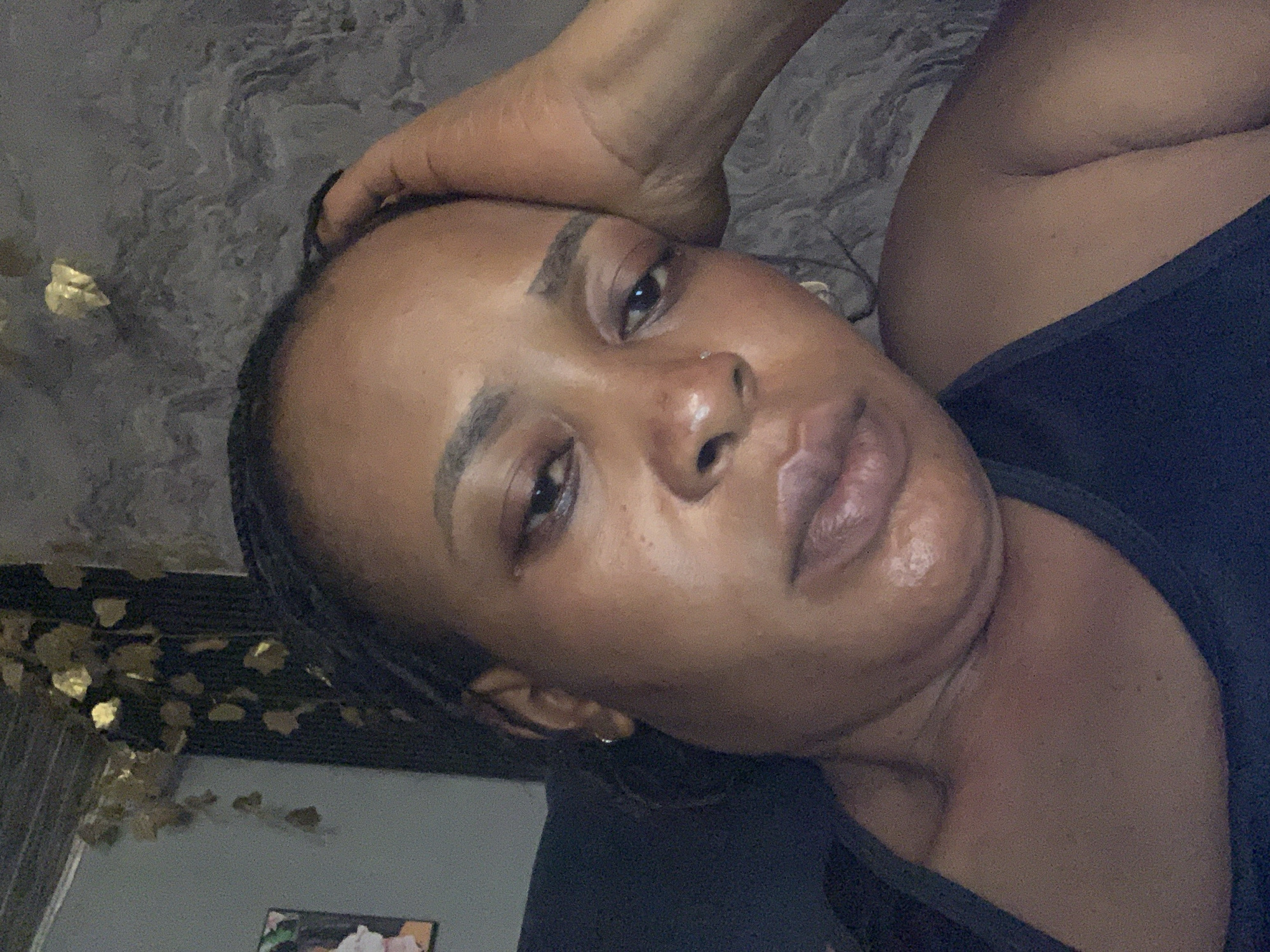 A close-up, head-and-shoulders shot captures a Black woman with braided hair, resting her right hand on her forehead. She is wearing a dark, possibly navy blue, sleeveless top. Her expression appears somber. The background is a textured wall with a gray, marbled pattern, and to the left, a dark vertical element with a gold, leafy garland draped over it is visible. The overall lighting is dim, suggesting an indoor setting. The image does not contain enough information to determine the location within Sabon Tasha, Nigeria.