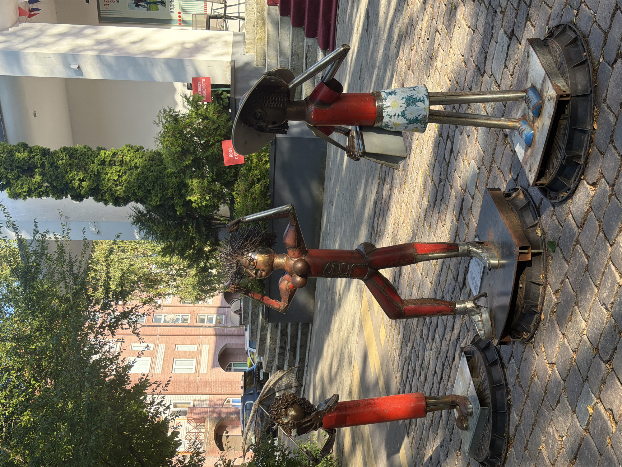 The image depicts three metal sculptures of figures standing on a cobblestone street in front of a building. The sculpture on the left is of a figure wearing a red dress and holding an umbrella. The central sculpture is of a figure with its arms raised, wearing a red top and pants. The sculpture on the right is of a figure wearing a red top, a patterned skirt, and a wide-brimmed hat, holding a bag. The background shows a building with red brick details, a white facade, and stairs leading up to an entrance. Two red signs are visible on the building's facade, with text in Cyrillic. The scene is illuminated by sunlight, casting shadows on the cobblestone. The location is identified as Zelenogradsk, Russia.