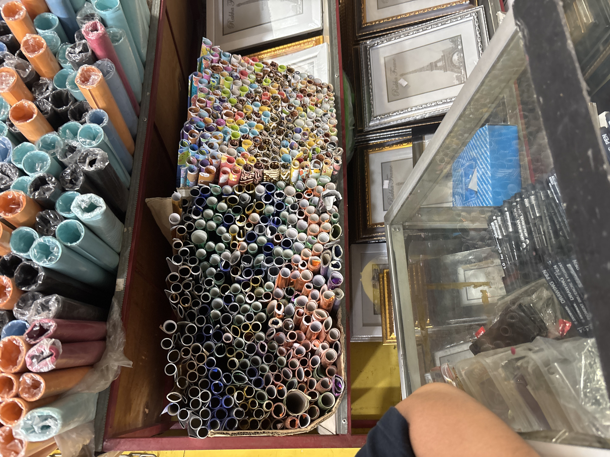 This image shows a shelf filled with rolls of paper or fabric in various colors, stacked vertically and horizontally. The rolls are of different sizes and patterns. Some are solid colors like orange, blue, green, and pink, while others have intricate designs. Below the rolls, there are framed pictures displayed, including one with an image of the Eiffel Tower. In the foreground, a glass display case contains various items, including pens and what appear to be art supplies. A person's arm and shoulder are visible on the left side of the image, suggesting they are interacting with the items in the display case. The scene appears to be a retail setting, possibly a craft or art supply store. The location is identified as Wanajaya, Indonesia.