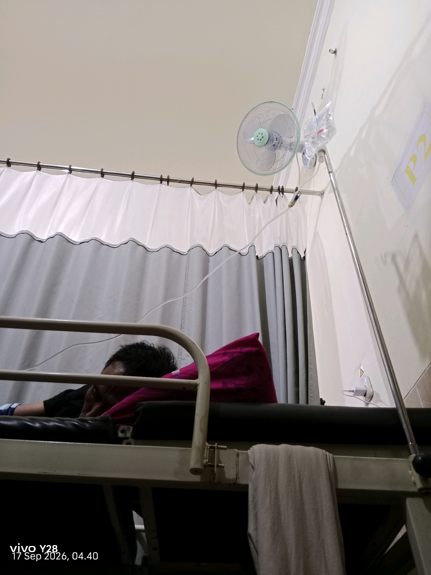 The image depicts an indoor scene, likely a hospital or medical facility. A person is lying on a bed, facing away from the camera, with their head resting on a pink pillow. The bed appears to be a simple metal frame. A curtain system, consisting of white and gray fabric panels, is suspended from a rod across the top of the scene, possibly serving as a privacy screen. To the right of the bed, an IV stand is visible, supporting a bag of intravenous fluid. A small electric fan is mounted on the wall above the bed. The lighting is dim, suggesting it might be nighttime or a poorly lit room. A watermark in the bottom left corner indicates the device used for capture ("vivo Y28") and the date and time ("17 Sep 2026, 04.40"). There is no direct indication of the location being Panggungan, Indonesia, as this information is not visually present in the image itself.