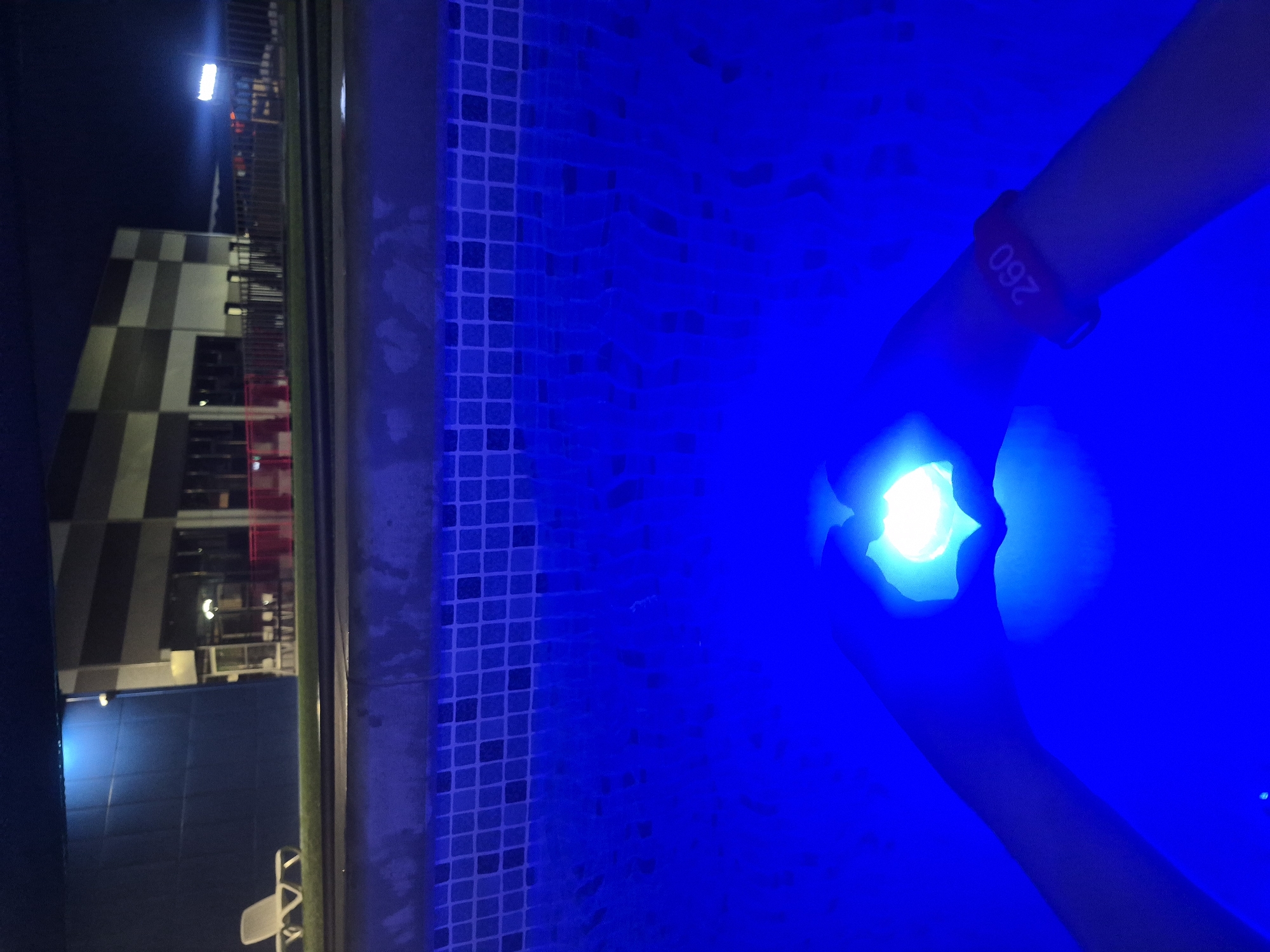 The image depicts a nighttime scene at an outdoor swimming pool in Votkinsk, Russia. The pool water is vividly illuminated by a strong blue light, casting a vibrant glow across the scene.
On the right side of the frame, a person's arm and hand are partially submerged in the glowing blue water. The hand is holding a brightly illuminated object, which emits a brilliant white or light blue light, serving as a focal point. A red wristband is worn on the person's arm, featuring the numbers "260" printed on it. The edge of the pool is lined with distinct blue and white mosaic tiles.
In the background, under a dark night sky, stands a modern building characterized by a checkered pattern of light and dark grey (or black) blocks. A fence or railing runs along the perimeter of the pool area, separating it from the background structures. Further back, a white outdoor chair is visible near the pool's edge. The scene is dominated by artificial lighting, highlighting the pool's deep blue and the bright object in the person's hand, suggesting a recreational or leisure activity taking place after sunset.