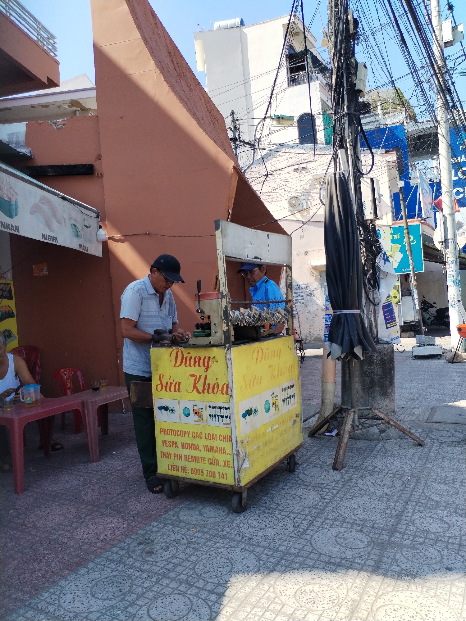 The scene shows two Vietnamese men operating a mobile key-cutting and lock repair service in the Bac Nha Trang Ward, Vietnam. The cart, labelled "Dùng Sửa Khóa," advertises locksmith services including key duplication for vehicles like Vespa, Honda, Yamaha, and remote key battery replacement, with a phone number listed. One man, wearing a cap and light blue shirt, is actively engaged in cutting keys using a machine mounted on the cart. The second man is also wearing a cap and a blue shirt, standing behind the key cutting machine. A customer sits at a red plastic table with drinks nearby. In the background, buildings and a complex tangle of power lines are visible.