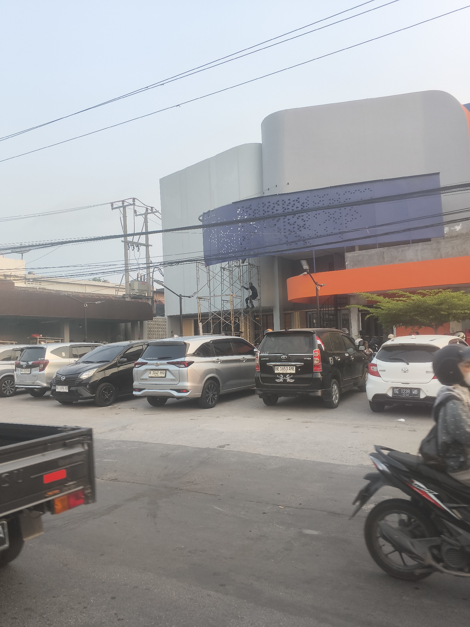 The image shows a street scene in Bandar Lampung, Indonesia, with a modern building under construction or renovation. In the foreground, several cars are parked, including a silver hatchback, a black sedan, and a silver SUV. A black pickup truck is partially visible on the left. A person on a motorcycle is riding past on the right side of the frame, with their face obscured by a helmet. The building in the background features a large, curved, light-colored facade and a lower section with orange and blue accents. Scaffolding is erected on the side of the building, indicating ongoing work, and a construction worker is visible on the scaffolding. Overhead, power lines and poles crisscross the sky. The sky is overcast with a pale, hazy light, suggesting it may be early morning or late afternoon.