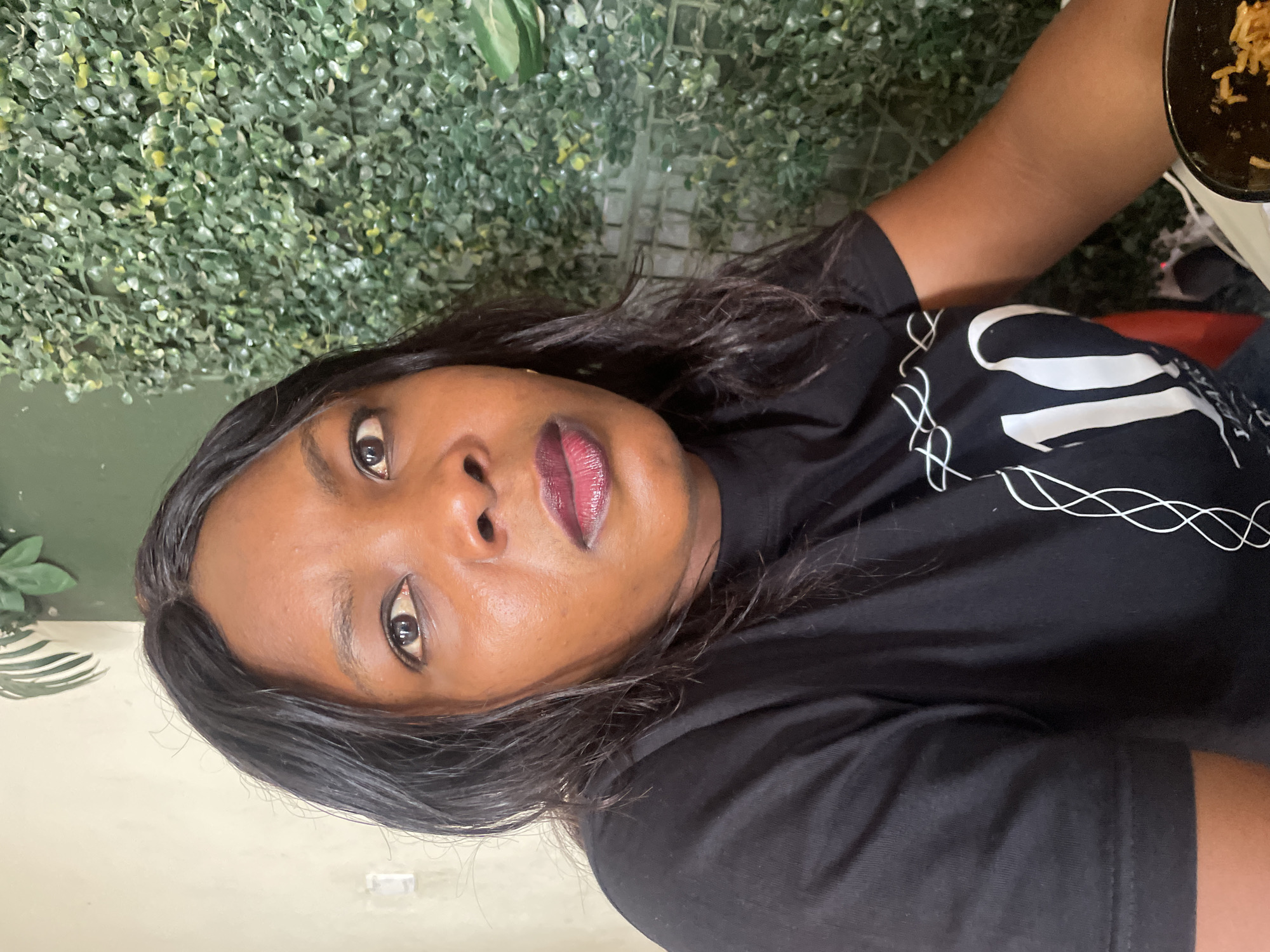 A medium-shot of a dark-skinned woman wearing a black t-shirt and dark lipstick. She has shoulder-length black hair. Part of a plate with food on it is visible in the upper right corner of the frame. Behind her is a wall covered in artificial greenery. The location is presumably Jos, Nigeria, based on the prompt.