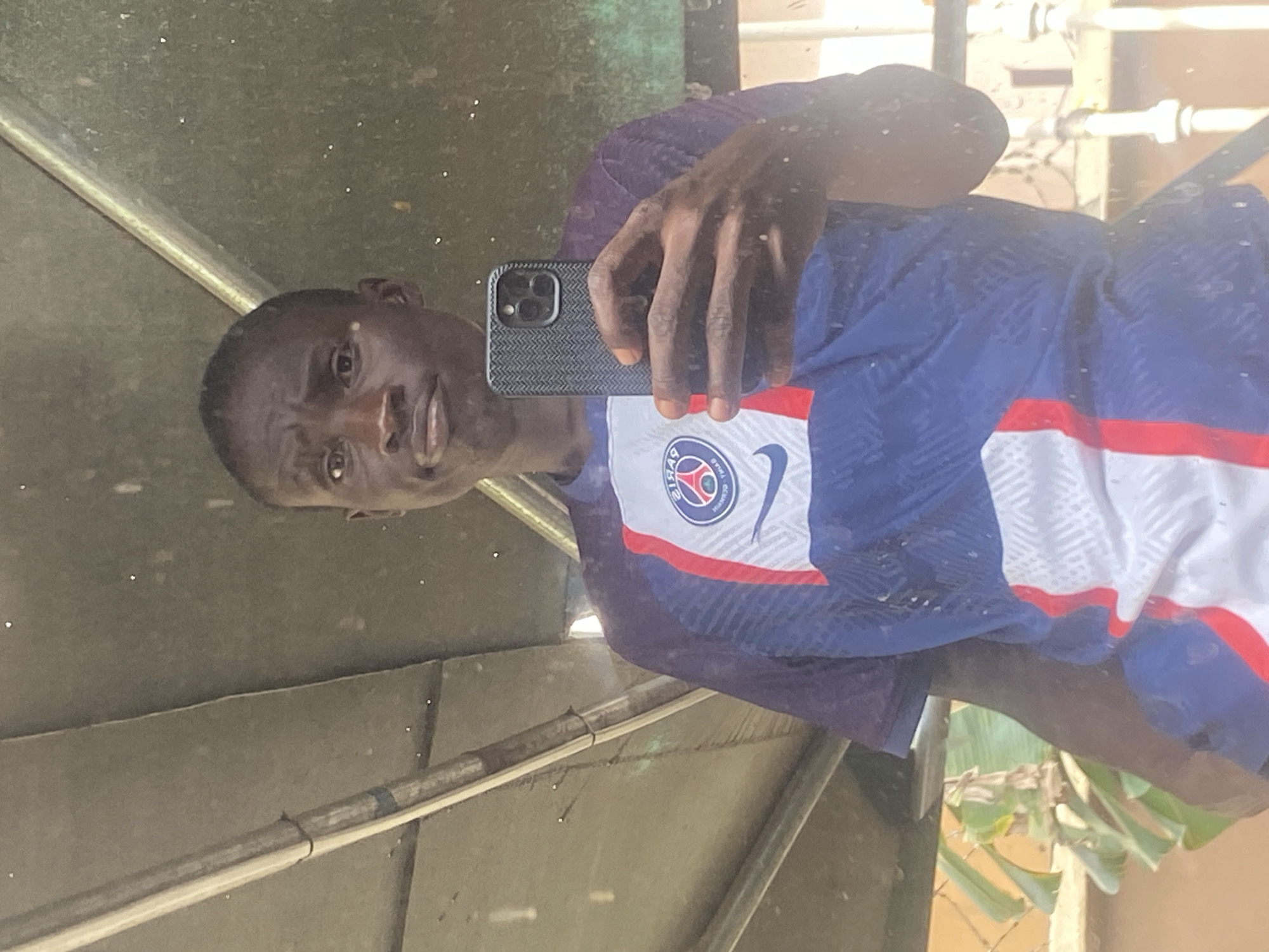 A dark-skinned person is visible in the image, apparently taking a selfie in a mirror. The person is wearing a blue, white and red jersey with the Paris Saint-Germain logo. The person is holding a phone in their hand. The location appears to be outdoors, potentially in Barden Yana, Nigeria, but the surroundings are ambiguous and lack specifics that could definitively confirm the location. The setting seems basic and lacks characteristics that would specify precise geographic details.