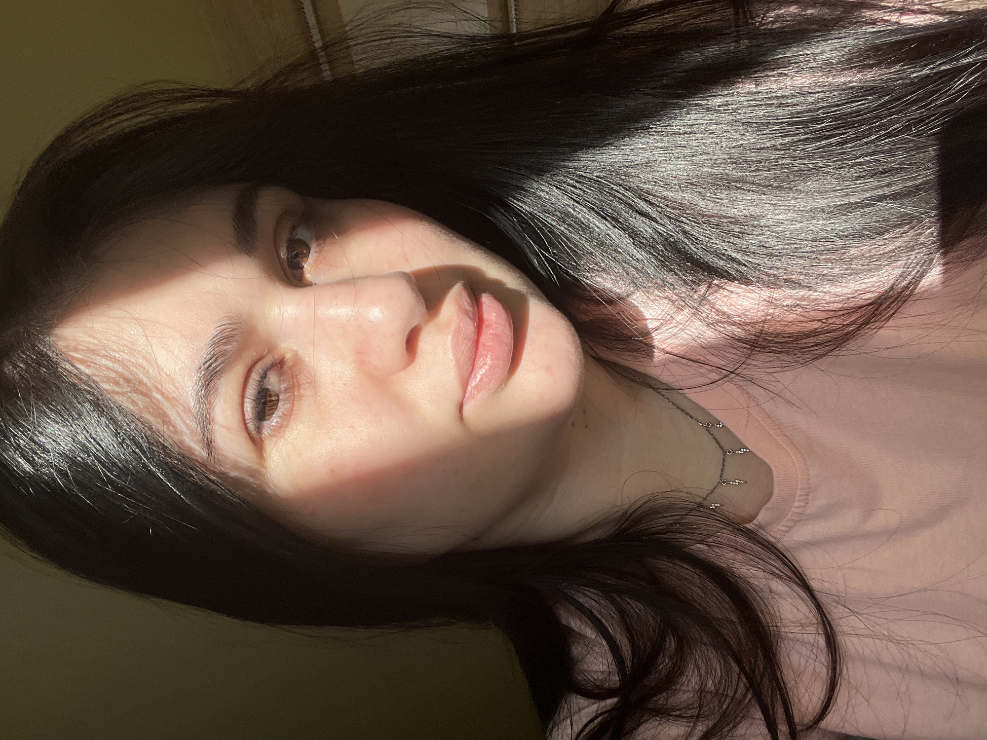 A close-up portrait shows a woman with dark hair wearing a pink shirt and a delicate silver necklace. The lighting is dramatic, with stark contrasts of bright sunlight and shadow across her face. Her expression is neutral as she looks towards the camera. The location is unspecified and there is no indication in this image of Chaikovsky, Russia.