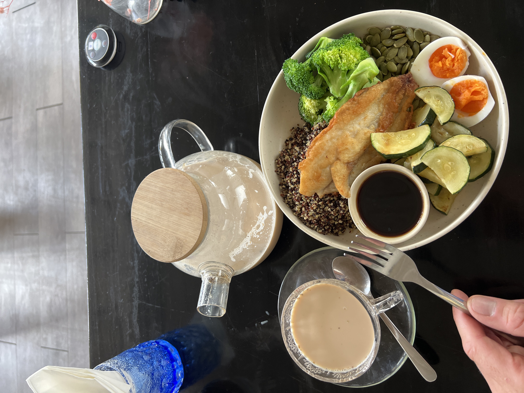An overhead, eye-level shot shows a meal and drink on a black, reflective table. In the center is a light-colored bowl containing a portion of quinoa, a piece of fried fish, broccoli florets, sliced zucchini, pumpkin seeds, and two halves of a hard-boiled egg with vibrant orange yolks. A small dish of dark sauce sits to the left of the bowl. To the left of the bowl, a clear glass teacup filled with a light brown liquid, likely coffee or tea, sits on a glass saucer. A spoon rests on the saucer. Behind the teacup is a clear glass teapot with a wooden lid, containing the same light brown liquid. A hand is visible in the bottom left corner, holding a silver fork over the teacup. To the far left, a blue glass is filled with white napkins. A small, round, black electronic device with colorful buttons is on the table in the upper right corner. The scene is set in North Kuta, Indonesia.