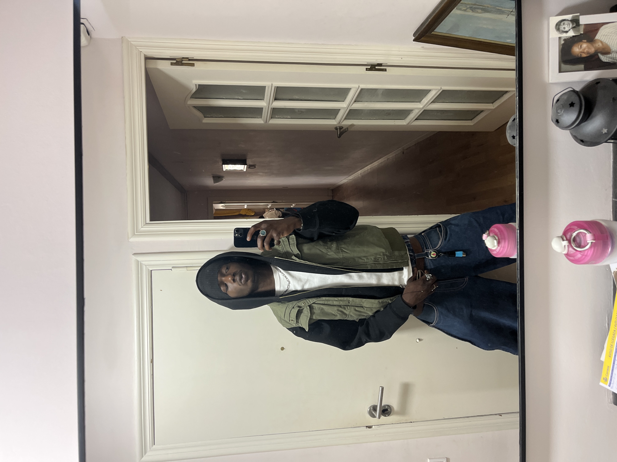 A man is taking a selfie in a mirror. He is wearing a black beanie, a white t-shirt, an olive green and black hooded jacket, and blue jeans. He is holding a black smartphone in his left hand. The mirror reflects a hallway with a white door and a door with frosted glass panes. On the wall next to the mirror, there is a framed photograph, a decorative wall lamp, and a pink soap dispenser. It is not possible to determine the location as Barcelona from the image alone.