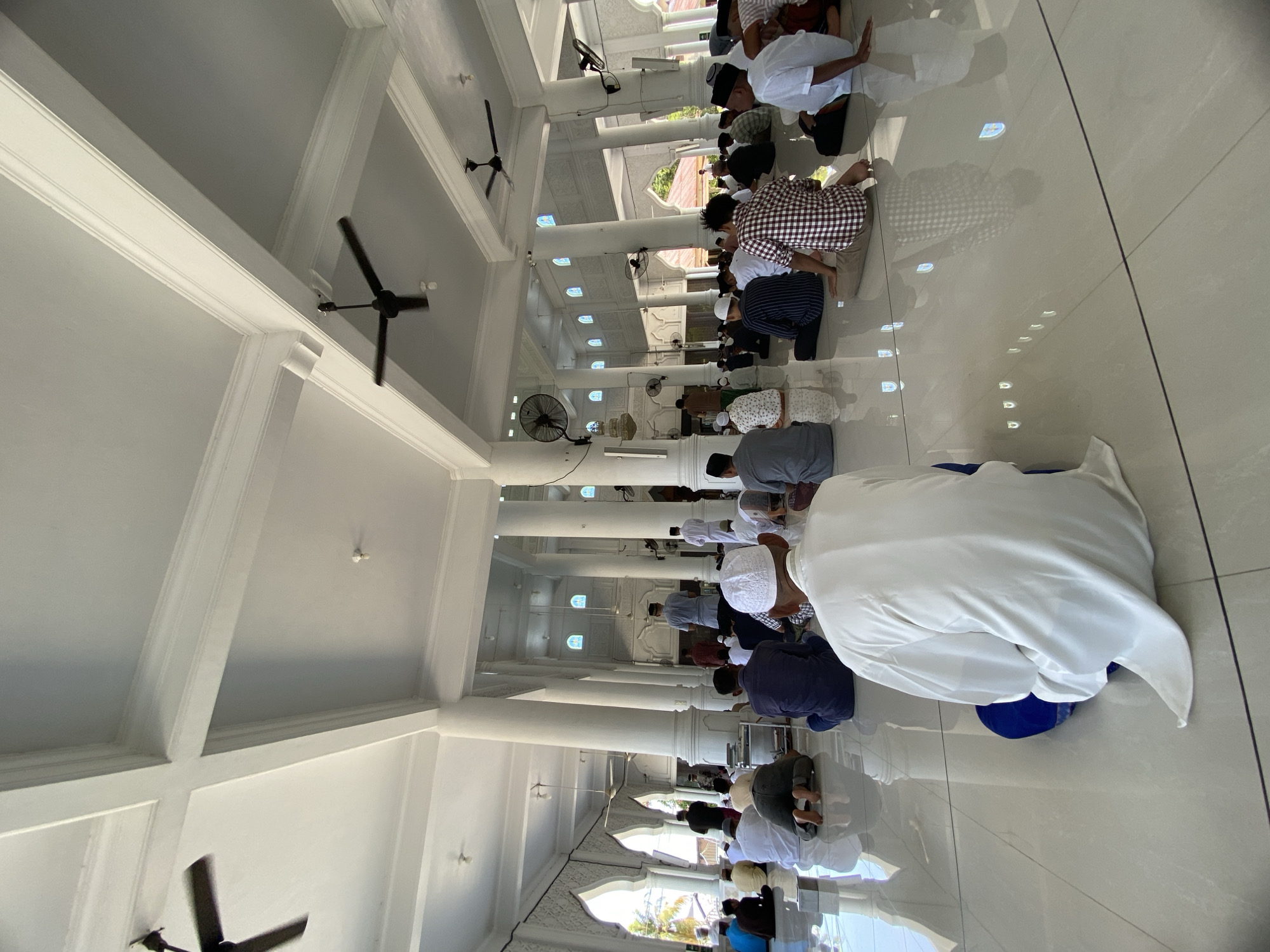 The image depicts a large group of people performing prayers inside what appears to be a mosque in Lhokseumawe, Indonesia. The individuals are kneeling in rows on a glossy tile floor. The architecture consists of white columns, a vaulted ceiling with multiple black ceiling fans, and arched windows. The setting is bright and well-lit.