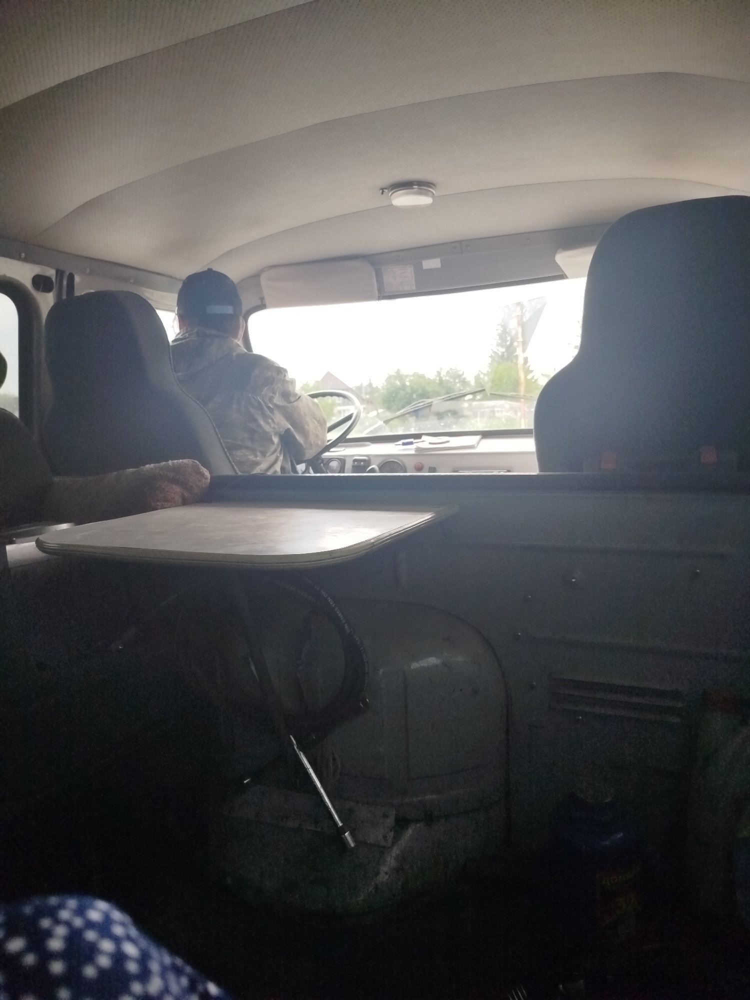 The image shows the interior of a vehicle, likely a van or truck, from the perspective of a passenger. A person wearing a camouflage jacket and a baseball cap is driving. The dashboard and steering wheel are visible, as is the windshield looking out onto a somewhat overcast day with trees and buildings in the distance. In the foreground, a tray table is mounted in front of the passenger seat, and part of a blue and white patterned blanket or clothing is visible in the lower left corner. A dark cylindrical object, possibly a tool or part of the vehicle's interior, is on the floor to the right. The environment outside appears to be rural or suburban. There are no specific indicators within the image to place it in NovoLybaevo, Russia.