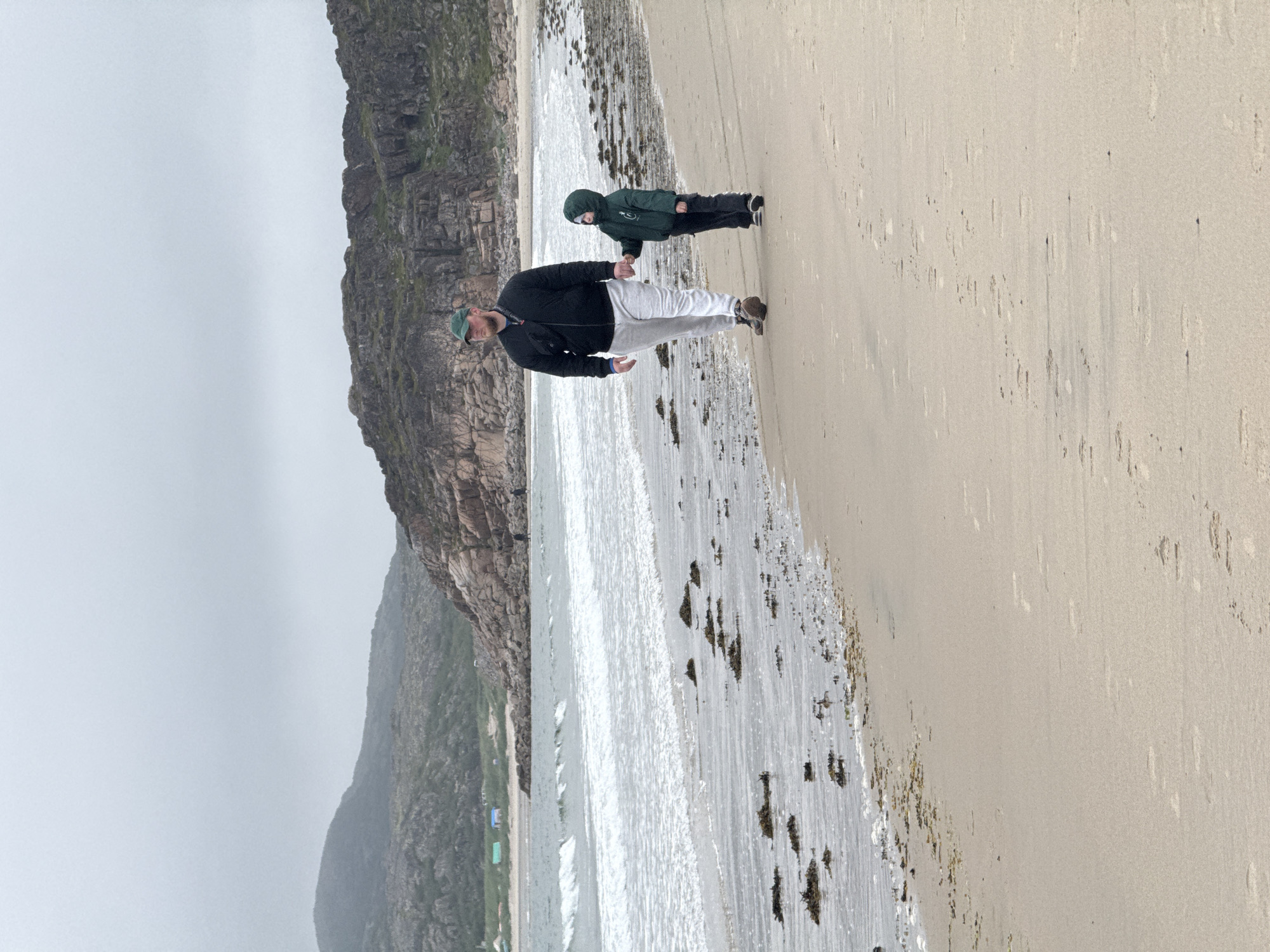 An adult male and a child are walking hand-in-hand on a sandy beach in Teriberka, Russia. The man is wearing a dark jacket, light-colored pants, and a green baseball cap. The child is wearing a dark green hooded jacket and dark pants. They are walking away from the viewer towards the left of the frame. The beach has light-colored sand with scattered seaweed and footprints. The ocean is to their left, with gentle waves rolling onto the shore. In the background, there are rocky hills and mountains under a cloudy sky. There are small structures visible in the distance along the coastline.
