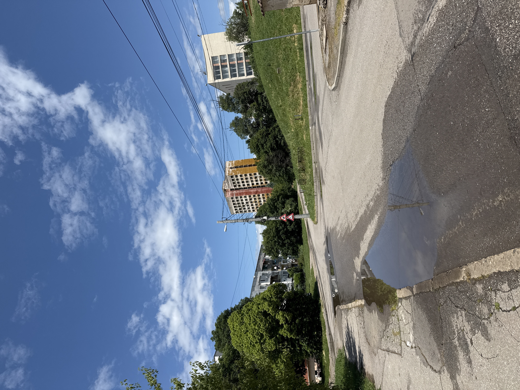 This image captures an outdoor urban landscape on a bright, sunny day with a clear blue sky dotted with scattered white clouds. The scene is likely set in Sofia, Bulgaria.
In the foreground, a paved asphalt road or path shows signs of wear with cracks and patches. A large, dark puddle on the road's surface acts as a natural mirror, reflecting the vibrant blue sky and fluffy white clouds above. A concrete curb delineates the road from a lush, green grassy bank that slopes gently upwards into the mid-ground.
Beyond the grassy incline, a variety of green trees and bushes are abundant, contributing to the verdant feel of the area. Several buildings rise into the background. To the left, a tall, light-colored building with multiple windows stands prominently. Further to the right, another multi-story structure appears to be under construction or undergoing renovation, characterized by its exposed concrete framework and some orange/yellow covering on its upper levels. In the distance, partly hidden by trees, a grey residential building with balconies and satellite dishes can be seen.
Multiple black utility lines crisscross the sky overhead. A utility pole with a streetlight arm is visible, and attached to it are two red-bordered, triangular warning road signs with white backgrounds. One sign features a black train engine symbol, indicating an unguarded railway crossing, while the other shows a double bend, first to the right.
The overall impression is one of a typical urban environment with a blend of natural elements and human-made structures, under the bright conditions of daytime. No people or specific activities are depicted.