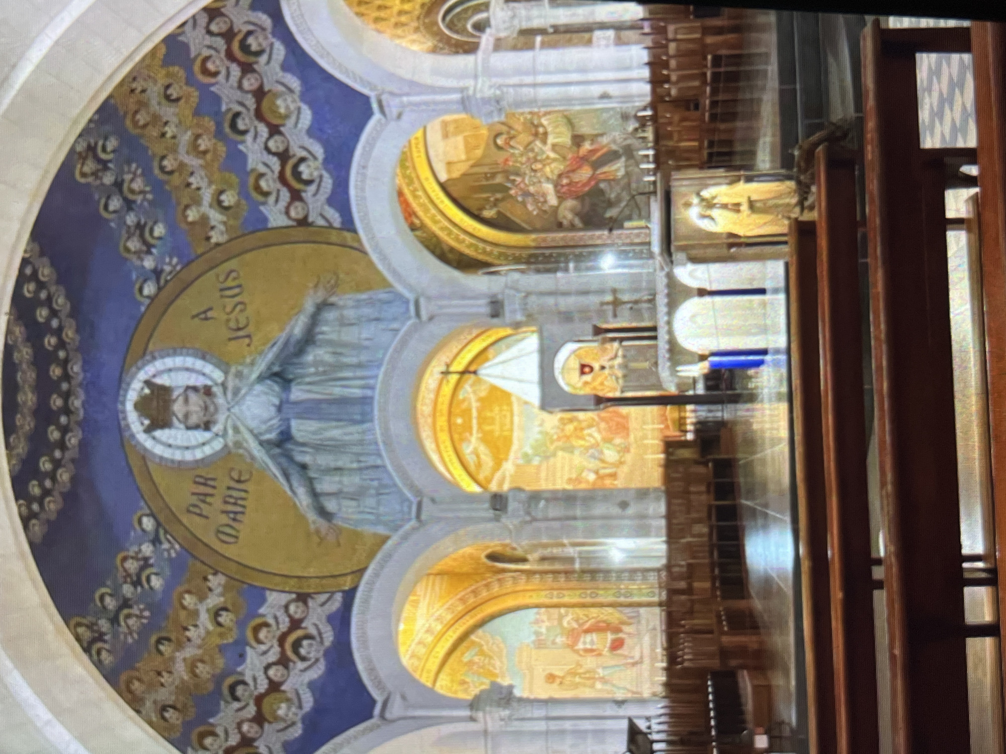 The image shows the interior of a basilica featuring a large semi-circular mosaic on the ceiling. The mosaic prominently displays a figure with arms outstretched, encircled by a golden halo, and the phrases "A JESUS" and "PAR MARIE." The surrounding area is decorated with rows of small, repeating figures, set against a blue background.
Below the mosaic, arches reveal further golden-toned murals. Wooden pews are visible in the foreground, suggesting the interior of a church. The basilica is located in the city of Tinaquillo, Venezuela.