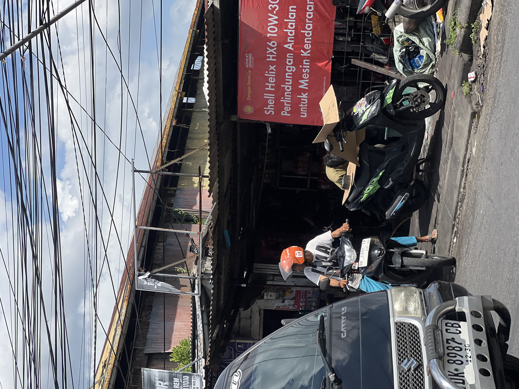 A medium shot captures a street scene in Denpasar, Indonesia, under a cloudy sky filled with a complex web of overhead wires. In the foreground, a black vehicle, identified by the text "CARRY 1.3" on its side, is partially visible on the left. Directly behind it, a person wearing an orange helmet and a white t-shirt rides a black motorcycle. The motorcycle has a license plate reading "DK 3395 TP". To the right of this motorcycle, another person, wearing a khaki shirt, is slumped over a black scooter, appearing to be resting or asleep. This scooter is adorned with green graphics and has a cardboard box secured to its back.
Behind the motorcycles, a row of dilapidated buildings with corrugated metal roofs lines the street. A large, red banner with white text is prominently displayed, advertising "Shell Helix HX6 10W-30" and describing its benefits for vehicle engines. The storefronts appear to be simple, open-air establishments. The street itself is paved, with a raised sidewalk or curb visible. Debris, including discarded cardboard and plastic bags, is scattered along the curb. The overall impression is one of a busy, perhaps somewhat weathered, urban environment.