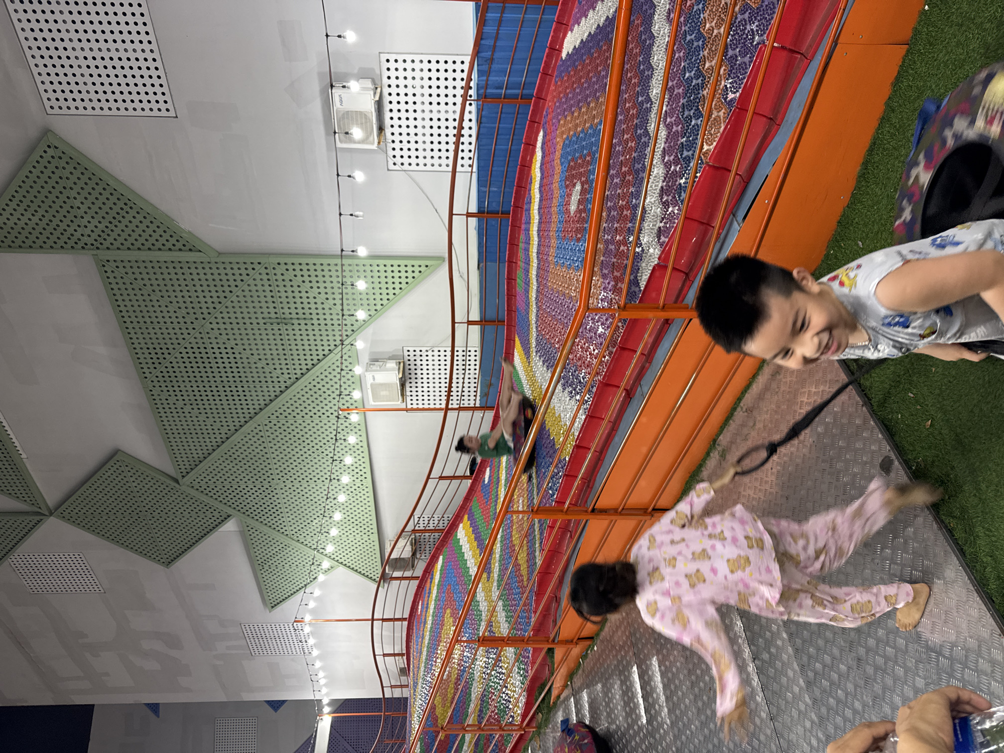 The scene is an indoor amusement park. The image shows children and adults enjoying the facilities. A boy in a grey shirt is smiling broadly. A child in pink pajamas is holding onto a rope. In the background, other people are either on or near a colorful, multi-tiered slide. The architecture of the building includes green perforated panels and square ventilation tiles on the ceiling. A line of small light bulbs runs along the ceiling. The location is likely in Đà Nẵng, Vietnam.