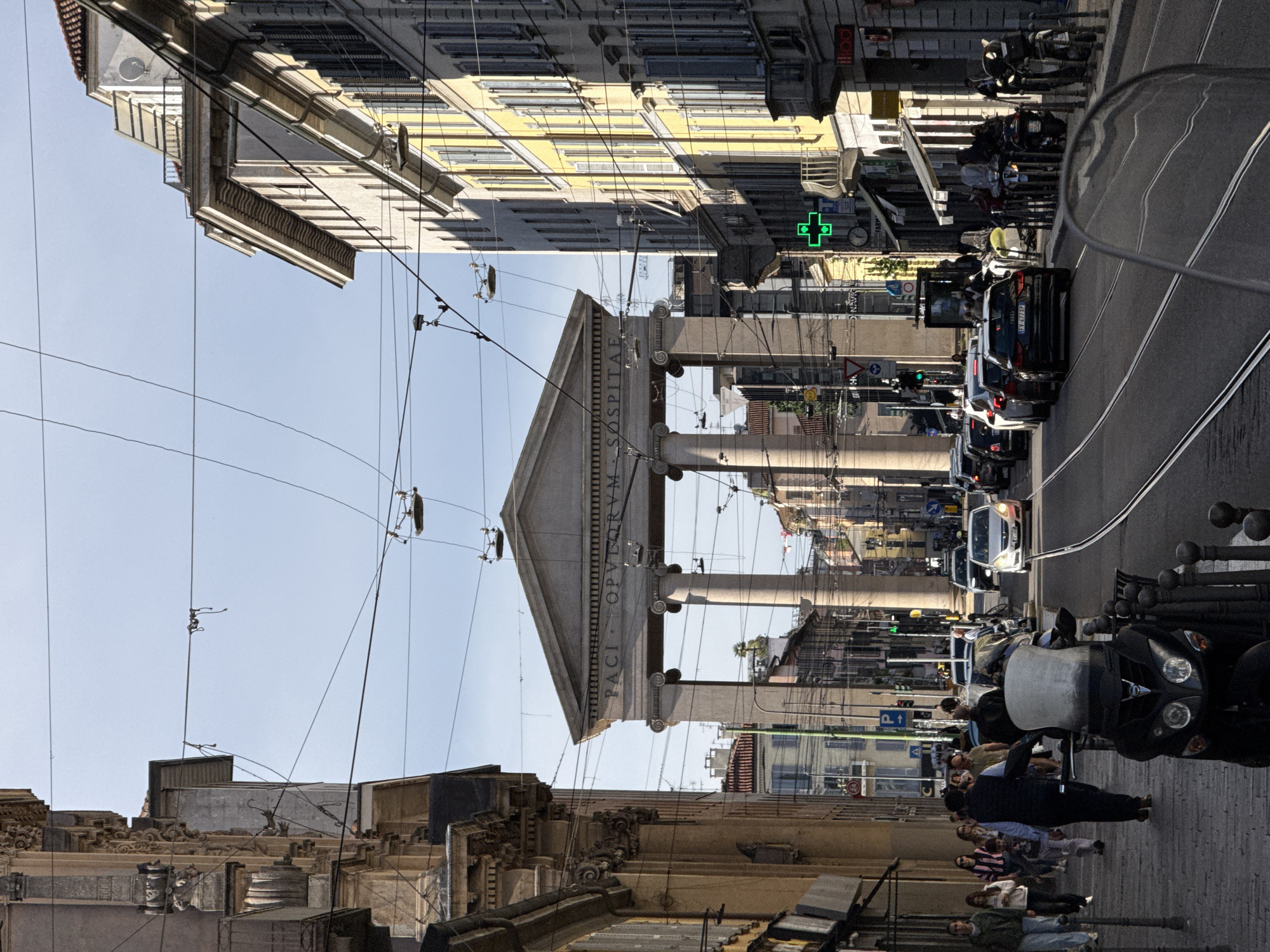 The image shows a street in Milan, Italy, featuring the neoclassic “Arco della Pace”. The archway is framed by surrounding buildings, with a bright, clear sky in the background. Overhead tram lines and streetlights are visible. The foreground depicts a sidewalk with pedestrians, as well as parked scooters, along with a line of cars. A pharmacy sign is visible along the buildings on the right side of the image.