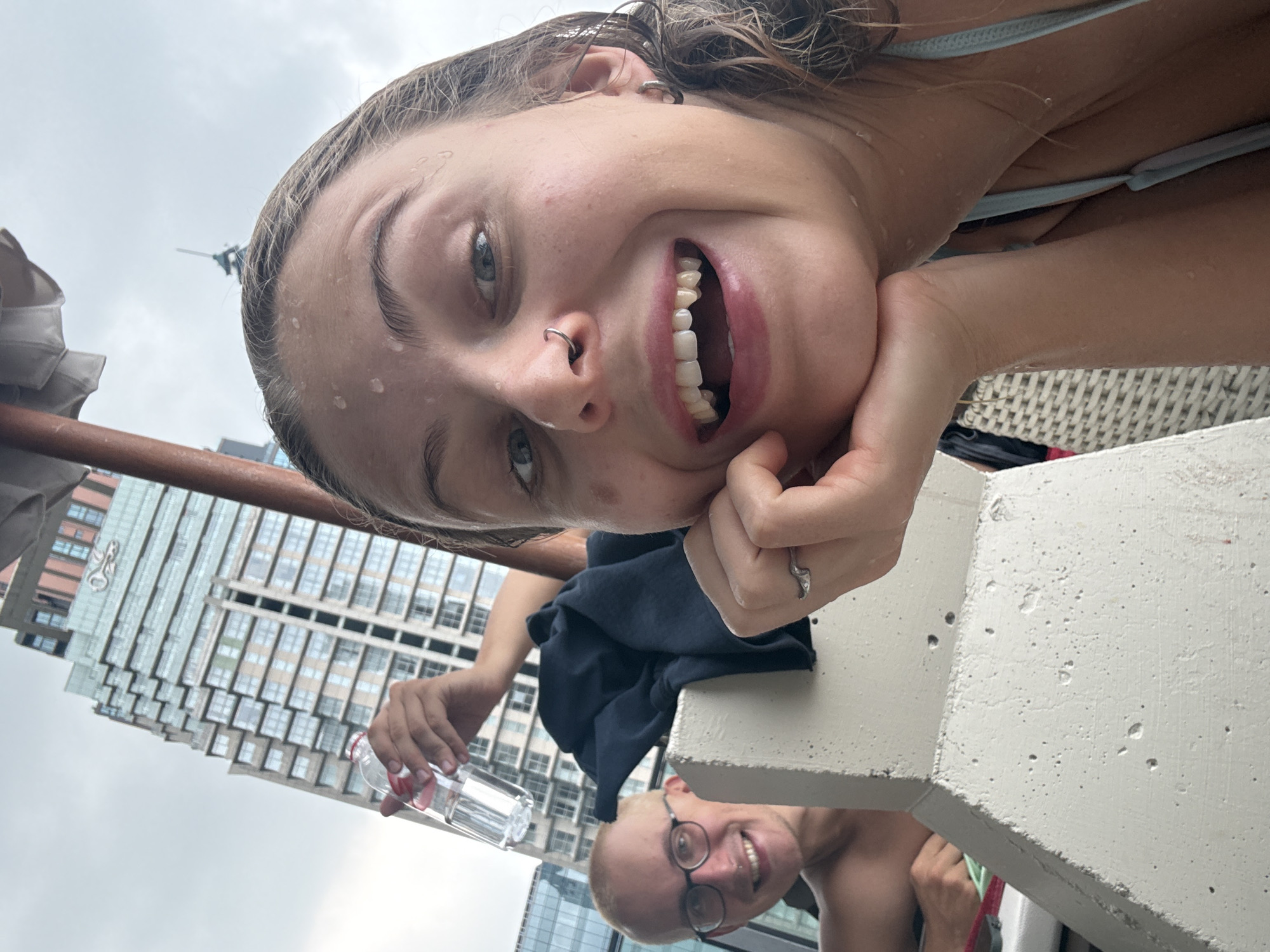 The image is a rotated selfie, capturing a close-up of a young woman smiling broadly under an overcast sky, with a city skyline in the background. The woman, positioned prominently, has wet, light brown hair and water droplets on her face, indicating she has been swimming or is near water. She wears a light blue swimsuit top, has a nose piercing, and a ring on her finger. Her expression is one of happiness and enjoyment.
In the background, a concrete ledge or barrier separates the foreground from the rest of the scene. Partially visible below the woman is another person, a young man with short blonde hair and glasses, also smiling. To the left, an arm holds a clear plastic water bottle. A dark blue fabric item is draped over the concrete. The backdrop features a tall, modern building with many windows, characteristic of an urban setting. The overall scene suggests an outdoor recreational area, possibly a rooftop pool or balcony in Bangkok, Thailand, given the location context. No visible text is present.