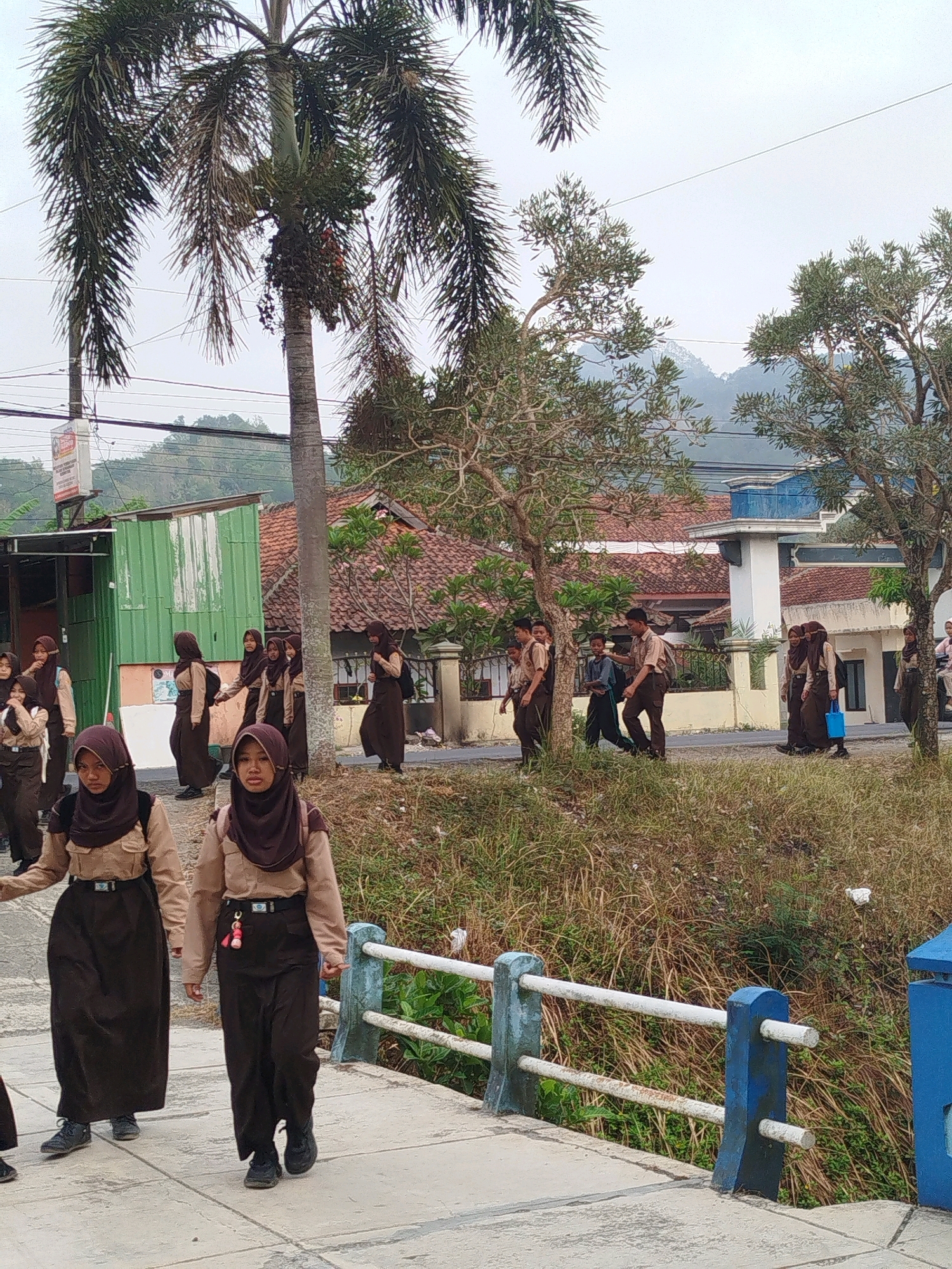 The image depicts a group of students in school uniforms walking along a paved path next to a grassy embankment. The students, predominantly female, wear brown headscarves and light brown shirts with brown skirts or trousers. Some carry backpacks. In the background, there are buildings with red-tiled roofs, palm trees, and a hilly landscape under an overcast sky. A blue and white railing runs along the side of the path. The scene appears to be in a semi-urban or rural setting.