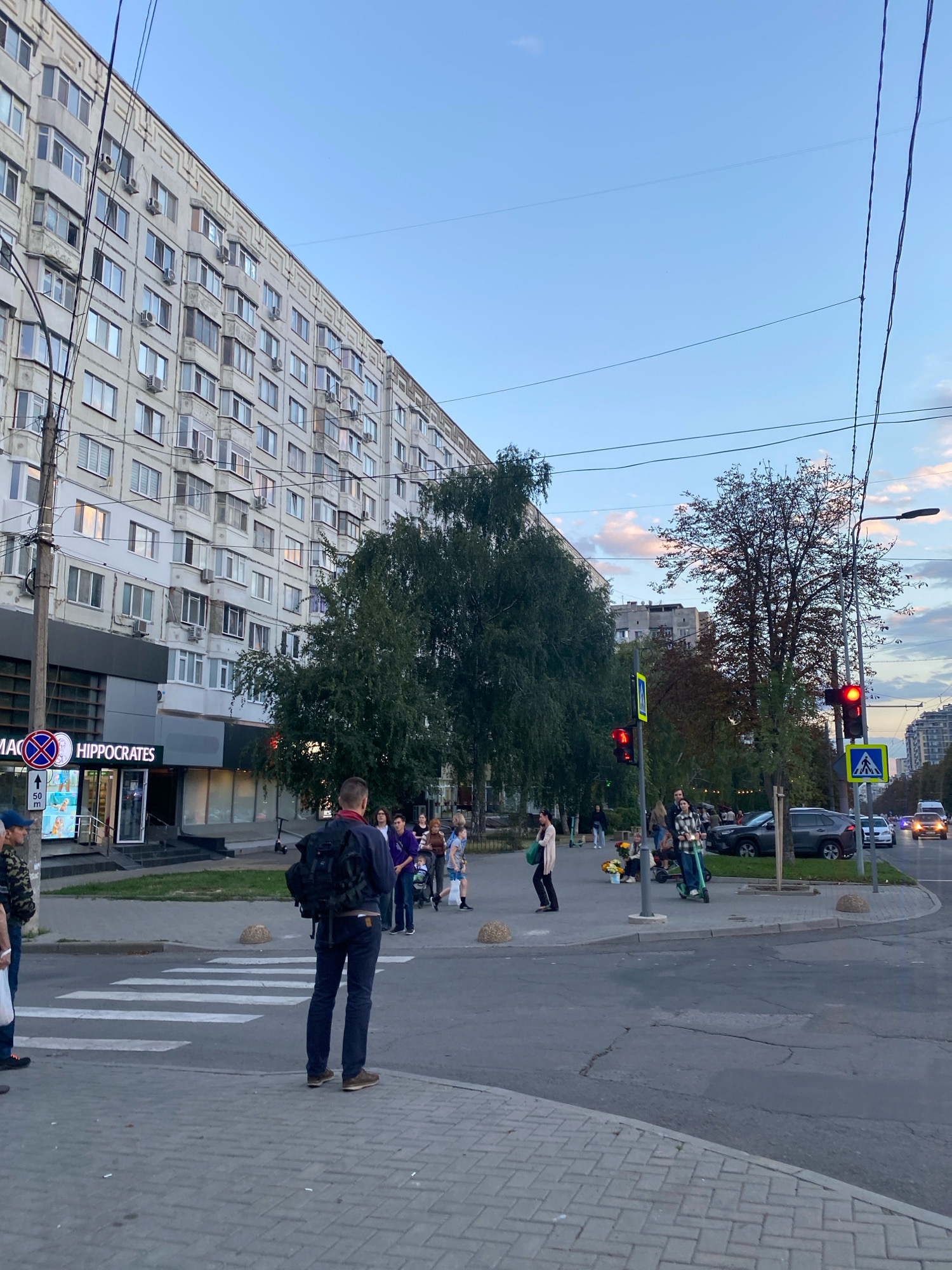 This image depicts a street scene in Chișinău, Moldova, likely during the late afternoon or early evening. The foreground shows a pedestrian crossing with a man wearing a dark backpack and jeans standing on the sidewalk facing away from the camera. In the middle ground, a large, leafy tree dominates the view, with several people gathered on the sidewalk near a building identified by the sign "HIPPOCRATES". Some individuals are on electric scooters, and a person is pushing a stroller. Traffic lights are visible, with one showing a red light. Across the street, cars are present, including a white van and other vehicles. The background features a long, multi-story apartment building with numerous windows and air conditioning units, characteristic of Soviet-era architecture. The sky is a pale blue with some wispy clouds, and power lines crisscross the upper portion of the image. The overall impression is a bustling urban environment with everyday street activity.